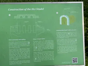 Đây là một tấm biển thông tin màu xanh lá cây với chữ viết màu trắng, được đặt ngoài trời tại khu di tích Thành nhà Hồ. Biển báo này giải thích về quá trình xây dựng Thành nhà Hồ bằng cả tiếng Việt và tiếng Anh.。Nội dung văn bản được ghi lại:。Tiêu đề chính (tiếng Anh): "Construction of the Ho Citadel"。Các chú thích trên sơ đồ (tiếng Việt):。- Vách tường đá。- Đất trộn sỏi。- Mặt đất。- Khối đá khóa đỉnh vòm。- Ụ đất tạo hình vòm。- Quét mã QR để khám phá thêm。Hoạt động được mô tả trên biển:。Tấm biển mô tả chi tiết các kỹ thuật và quy trình xây dựng Thành nhà Hồ, bao gồm ba giai đoạn chính:。1.  **Khai thác và chế tác đá (Stone quarrying and crafting):** Đá được khai thác từ các mỏ đá vôi ở núi An Tôn và Xuân Đài, sau đó được chế tác thành các khối hình chữ nhật hoặc hình vuông.。2.  **Vận chuyển đá (Stone transportation):** Các khối đá được vận chuyển bằng đường bộ sử dụng hệ thống "băng chuyền" với các khúc gỗ và bi đá, hoặc vận chuyển bằng bè tre trên sông Mã đến Bến Đá.。3.  **Xây dựng tường thành và cổng thành (Constructing citadel walls and gates):** Các khối đá nặng hàng chục tấn được đưa lên cao bằng các đường dốc bằng đất. Các cổng vòm được xây dựng bằng cách xếp các khối đá hình nêm xung quanh một ụ đất làm lõi định hình.。Các vật thể được hiển thị:。- Một biển báo thông tin có khung kim loại.。- Các sơ đồ kỹ thuật minh họa cách xây tường, làm đường dốc và xây cổng vòm.。- Một mã QR ở góc dưới bên phải để khách tham quan tìm hiểu thêm thông tin.。（日本語訳）。これは、胡朝の城跡の屋外に設置された、白い文字が書かれた緑色の案内板です。この看板は、胡朝の城の建設過程について、ベトナム語と英語の両方で説明しています。記載されているテキストの内容：。主な見出し（英語）：「Construction of the Ho Citadel」。図上の各注釈（ベトナム語）：。- 石壁。- 砂利混じりの土。- 地面。- アーチの頂点を固定する石材。- アーチを形成するための土の盛り土。- QRコードをスキャンしてさらに詳しく。看板に記述されている活動：。看板は、胡朝の城の建設技術と工程を詳細に記述しており、主に以下の3つの段階を含みます：。1.  **石の採掘と加工 (Stone quarrying and crafting):** 石はアントン山とスアンダイ山の石灰岩採石場から採掘され、その後、長方形または正方形のブロックに加工されました。2.  **石の運搬 (Stone transportation):** 石のブロックは、丸太と石の玉を使った「コンベア」システムを利用して陸路で運ばれるか、またはマー川を竹のいかだでベンダー（石の船着き場）まで運ばれました。3.  **城壁と城門の建設 (Constructing citadel walls and gates):** 数十トンの重さの石ブロックは、土の坂道を使って高い所へ持ち上げられました。アーチ門は、成形用の芯となる土の盛り土の周りに楔形の石ブロックを積み重ねることによって建設されました。表示されている物体：。- 金属フレーム付きの案内板。- 壁の建設方法、坂道の作り方、アーチ門の建設方法を説明する技術的な図解。- 観光客がさらに情報を得るための、右下隅にあるQRコード。