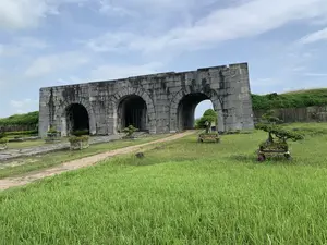 Bức ảnh chụp một công trình kiến trúc cổ, đó là một cổng thành lớn được xây bằng những khối đá khổng lồ. Cổng thành có ba cửa ra vào hình vòm. Đây là một di tích lịch sử, có khả năng là cổng thành của Thành nhà Hồ tại Thanh Hóa. Phía trước cổng là một bãi cỏ rộng lớn, xanh mướt, có một con đường lát đá dẫn vào trong. Một vài chậu cây cảnh bonsai được đặt trên bãi cỏ và dọc theo lối đi. Phía sau và bên cạnh cổng thành là các bờ tường đất phủ đầy cỏ. Bầu trời có mây. Không có người và không có hoạt động nào đang diễn ra. Không có bất kỳ văn bản hay chữ viết nào trong ảnh.。（日本語訳）。写真は古い建築物を写しており、それは巨大な石のブロックで建てられた大きな城門です。城門には3つのアーチ型の出入り口があります。これは歴史的遺跡であり、タインホア省にある胡朝の城の城門である可能性があります。門の前には広大で青々とした芝生が広がり、中へと続く石畳の道があります。いくつかの盆栽の鉢植えが芝生の上と通路に沿って置かれています。城門の後ろと横には、草で覆われた土の壁があります。空には雲があります。人はおらず、何の活動も行われていません。写真にはいかなるテキストや文字もありません。