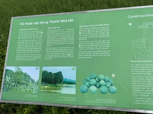 Bức ảnh chụp cận cảnh một bảng thông tin màu xanh lá cây có khung kim loại, đặt ngoài trời trên nền cỏ. Bảng thông tin này cung cấp chi tiết về các kỹ thuật xây dựng Thành Nhà Hồ bằng cả tiếng Việt và tiếng Anh, kèm theo hình ảnh và sơ đồ minh họa. Nội dung văn bản tiếng Việt trên bảng được ghi lại đầy đủ như sau:。**Kỹ thuật xây dựng Thành Nhà Hồ**。**Khai thác và chế tác đá**。Đá được khai thác và gia công tại chỗ từ các núi đá vôi như An Tôn, Xuân Đài ①, v.v. cách đây khoảng 2 đến 4 km. Đá ở đây có cấu tạo tự nhiên từng thớ chồng xếp, rất thuận lợi để bóc tách và chế tác.。Từ núi cao, người thợ xưa chọn những thớ đá lớn, “nạc” để khoan, đục, tạo ra các rãnh lỗ ở giữa các thớ và dùng các công cụ thủ công như búa, nêm, chét, đòn bẩy, v.v. để tách từng khối nhỏ và đục đẽo thành khối vuông hoặc chữ nhật. Sau đó, chúng được làm phẳng để giảm trọng lượng và dễ dàng vận chuyển về thành.。Tại chân tường thành, những người thợ đá tiếp tục gia công, chế tác để tạo ra các hình khối theo ý muốn, đảm bảo thẩm mỹ trước khi đưa lên xây dựng thành.。**Vận chuyển đá**。Để vận chuyển đường bộ, những người thợ đã đắp một con đường vững chắc. Sau đó, họ dùng sức người, sức kéo của trâu, voi để trượt các tảng đá trên một “băng chuyền” thủ công làm bằng các trục gỗ và con lăn đá tròn ② xếp cách đều nhau. Để khối đá không bị dịch khỏi băng chuyền, dân phu dùng đòn bẩy ở hai bên điều chỉnh các khối đá dịch chuyển đúng hướng.。Về đường thủy, có thể sông Mã và vùng đầm lầy phía Bắc Hoàng thành cũng là tuyến vận chuyển đá. Đá được chuyển từ núi An Tôn xuống sông, đưa lên bè mảng và xuôi dòng về khu vực Bến Đá ③. Sau đó, đá được vận chuyển bằng đường bộ vào thành.。**Xây dựng tường và cổng thành**。Để đưa những phiến đá nặng hàng chục tấn lên xây lắp tường thành, cổng thành ④, những người thợ xây thành đã đắp đất tạo nên một con dốc nghiêng chắc chắn. Thành xây đến đâu, con dốc được tôn cao đến đó và kéo dài ra để dễ dàng đưa đá lên.。Để xây được các vòm cửa bằng đá, người ta đã cho đắp ụ đất hình vòm cửa ⑤, sau đó dùng đá đã được chế tác hình thang cân ghép lên trên. Sau khi ghép xong, họ moi lõi đất ra để tạo vòm cửa.。Tường thành được xây bằng đá xếp theo hình chữ công (工). Ở mặt trong, đá được chèn nối tiếp theo kiểu cài nanh sấu giúp tường vững chắc.。Các hình ảnh minh họa trên bảng bao gồm:。1.  Ảnh chụp 'Các lớp đá vôi trên núi Xuân Đài'.。2.  Ảnh chụp 'Con lăn đá được tìm thấy tại các di tích và trong cộng đồng cư dân quanh thành'.。3.  Ảnh chụp 'Bến Đá được cho là nơi tập kết đá', cho thấy một khúc sông và vài chiếc bè hoặc thuyền nhỏ.。4.  Sơ đồ kỹ thuật xây dựng tường thành.。5.  Sơ đồ kỹ thuật xây vòm cửa.。（日本語訳）。この案内板は、胡朝の城の建設技術について、ベトナム語と英語の両方で詳細を説明しており、イラスト画像や図解も添えられている。案内板に記載されているベトナム語のテキスト内容は以下の通りである。**胡朝の城の建設技術**。**石材の採掘と加工**。石材は、約2～4km離れたアン・トン山、スアン・ダイ山①などの石灰岩の山からその場で採掘・加工された。ここの石は、層が積み重なった自然な構造をしており、剥離や加工に非常に都合が良かった。高い山から、昔の職人たちは大きくて「肉厚な」石の層を選び、ドリルで穴を開け、鑿（のみ）で彫り、層の間に溝を作り、ハンマー、くさび、のみ、てこなどの手工具を使って小さな塊に分離し、正方形または長方形のブロックに削り出した。その後、重量を減らし、城への輸送を容易にするために平らにされた。城壁の麓では、石工たちがさらに加工・製作を続け、望み通りの形状を作り出し、城の建設に使用する前に美観を確保した。**石材の輸送**。陸上輸送のために、職人たちは頑丈な道を築いた。その後、人力、水牛や象の牽引力を利用して、等間隔に並べられた木製の軸と丸い石のローラー②で作られた手動の「コンベア」の上で石の塊を滑らせた。石の塊がコンベアからずれないように、人夫たちは両側でてこを使い、石の塊が正しい方向に移動するように調整した。水上輸送については、マー川と皇城北部の湿地帯も石材の輸送路であった可能性がある。石材はアン・トン山から川に下ろされ、いかだに乗せられて川を下り、ベン・ダー（石の船着き場）③地区へと運ばれた。その後、石材は陸路で城内に運ばれた。**城壁と城門の建設**。数十トンの重さがある石の板を城壁や城門④の建設のために持ち上げるため、城の建設者たちは土を盛って頑丈な傾斜路を作った。城の建設が進むにつれて、傾斜路も高くされ、石を運び上げやすいように長くされた。石造りのアーチ門を建設するために、人々はアーチ門の形をした土の塚⑤を築き、その上に等脚台形に加工された石を組み上げた。組み上げが終わった後、土の芯を掘り出してアーチ門を形成した。城壁は「工」の字の形に石を積んで建てられた。内側では、石は「ワニの歯」のように交互に差し込む形で連結され、壁を頑丈にしている。案内板のイラスト画像には以下が含まれる。1.  「スアン・ダイ山の石灰岩層」の写真。2.  「遺跡や城周辺の住民コミュニティで発見された石のローラー」の写真。3.  「石の集積所であったとされるベン・ダー」の写真で、川の一部と数艘のいかだまたは小舟が写っている。4.  城壁の建設技術の図解。5.  アーチ門の建設技術の図解。