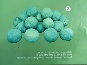 Đây là một bảng thông tin có nền màu xanh lá cây, có thể là tại một bảo tàng hoặc khu di tích. Trên bảng là hình ảnh chụp một đống con lăn đá hình cầu, có màu xanh xám. Văn bản trên bảng được ghi lại đầy đủ như sau: Phía trên bên phải có số '2' trong vòng tròn trắng. Phía dưới là dòng chú thích bằng tiếng Việt 'Con lăn đá được tìm thấy tại các di tích và trong cộng đồng cư dân quanh thành' và dòng chú thích tương ứng bằng tiếng Anh 'Stone balls found in temples and communities around the citadel'.。（日本語訳）。これは緑色の背景の案内板で、博物館か史跡にあるものかもしれません。板には、青灰色の球形をした石のローラーの山を写した写真があります。板の文言は、以下のように完全に記録されています。右上には、白い丸の中に数字の「2」があります。その下には、ベトナム語のキャプション「Con lăn đá được tìm thấy tại các di tích và trong cộng đồng cư dân quanh thành」と、対応する英語のキャプション「Stone balls found in temples and communities around the citadel」があります。