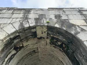 Bức ảnh chụp cận cảnh một vòm cổng lớn bằng đá, nhìn từ dưới lên. Đây là một phần của một công trình kiến trúc cổ, có khả năng là cổng của một thành cổ, cụ thể là Thành nhà Hồ dựa trên thông tin địa lý. Vòm cổng được xây dựng kiên cố từ những phiến đá lớn, hình chữ nhật, xếp chồng lên nhau. Bề mặt đá đã cũ, có nhiều vết ố đen và rêu, cây cỏ dại mọc trong các kẽ hở, cho thấy dấu ấn của thời gian. Không có chữ viết hay biển hiệu nào trong ảnh. Phía trên cùng của ảnh là bầu trời với những đám mây.。（日本語訳）。石造りの大きなアーチ門を、下から見上げたクローズアップ写真。これは古い建築物の一部で、地理情報に基づくと、おそらく城塞の門、具体的には胡朝の城の門である。アーチ門は、長方形の大きな石板を積み重ねて頑丈に造られている。石の表面は古く、黒いシミや苔が多く、隙間には雑草が生えており、時の流れを感じさせる。写真には文字や看板はない。写真の最上部は雲のある空である。