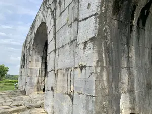 Hình ảnh chụp một góc của một công trình kiến trúc bằng đá cổ kính, có khả năng là một phần của Thành nhà Hồ. Công trình là một bức tường thành khổng lồ được xây dựng từ những khối đá lớn, hình chữ nhật, xếp chồng lên nhau một cách vững chãi. Bức tường có các cổng vòm lớn. Bề mặt đá đã bị phong hóa nặng theo thời gian, thể hiện qua các vết nứt, những mảng màu sẫm do ẩm mốc và thời tiết. Nền đất phía trước chân tường được lát bằng những phiến đá không đều nhau. Phía bên trái có cây cối và bãi cỏ. Bầu trời phía trên có nhiều mây. Không có người hay hoạt động nào diễn ra trong ảnh. Có một biển báo nhỏ ở phía xa bên trái nhưng không thể đọc được nội dung.。（日本語訳）。古い石造りの建築物の一角を撮影した写真で、おそらく胡朝の城の一部である。この建造物は、大きな長方形の石のブロックを頑丈に積み重ねて造られた巨大な城壁である。城壁には大きなアーチ状の門がある。石の表面は時間とともにひどく風化しており、ひび割れや、湿気や天候による黒ずんだ部分にそれが現れている。城壁の麓の前の地面は、不揃いな石板で舗装されている。左側には木々や芝生がある。上空の空は雲が多い。写真には人や活動は見られない。左奥に小さな看板があるが、内容は読み取れない。