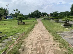 Bối cảnh là một không gian rộng lớn, có thể là một công viên hoặc khu di tích lịch sử, với một con đường lát gạch đỏ chạy giữa bãi cỏ xanh. Trên ki-ốt màu xanh lam ở phía xa bên trái có dòng chữ 'NƠI GIỚI THIỆU VÀ BÁN VÉ'. Hai bên con đường có đặt các chậu cây cảnh bonsai được trang trí công phu. Trong khung cảnh, có hai người phụ nữ đội nón lá đang đi xe đạp và một vài du khách khác đang đi bộ ở phía xa. Xung quanh có nhiều cây xanh, cột đèn và một số lá cờ hội đang được treo, tạo nên một không gian yên tĩnh và mang đậm nét văn hóa.。（日本語訳）。背景は広大な空間で、公園あるいは史跡のようであり、緑の芝生の真ん中を赤レンガ敷きの道が走っています。左奥にある青いキオスクには「NƠI GIỚI THIỆU VÀ BÁN VÉ」という文字があります。道の両側には、手の込んだ装飾が施された盆栽の鉢植えが置かれています。風景の中には、ノンラーをかぶった二人の女性が自転車に乗っており、遠くには他の観光客が数人歩いています。周囲には多くの緑の木々、街灯、そして祭りの旗がいくつか掲げられており、静かで文化的な趣の濃い空間を創り出しています。