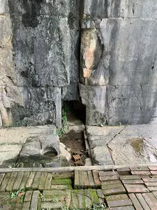 Bức ảnh chụp cận cảnh một phần của một bức tường đá cổ, có thể là một phần của một thành cổ hoặc di tích lịch sử. Bức tường được xây dựng từ những khối đá lớn, xếp chồng lên nhau. Bề mặt đá đã cũ, nhuốm màu thời gian với những mảng sẫm màu do ẩm và phong hóa. Tại chân tường có một lỗ hổng nhỏ, tối, bên trong có cây cỏ mọc. Nền đất phía trước tường không bằng phẳng, bao gồm các phiến đá, một vũng nước nhỏ, và vài viên gạch vỡ. Ở gần nhất trong ảnh là một lối đi lát bằng gạch đỏ cũ kỹ, phủ đầy rêu xanh và cỏ dại mọc xen kẽ giữa các kẽ gạch. Không có người hay hoạt động nào được ghi lại. Toàn bộ khung cảnh toát lên vẻ cổ kính, ẩm ướt và tĩnh lặng, đặc trưng của một di tích lịch sử.。（日本語訳）。古い石壁の一部を接写した写真で、城壁か史跡の一部である可能性があります。壁は大きな石のブロックを積み重ねて造られています。石の表面は古く、湿気や風化による黒ずんだ部分があり、時の流れを感じさせます。壁の根元には、暗く小さな穴があり、その中には草木が生えています。壁の前の地面は平らではなく、石板、小さな水たまり、そしていくつかの割れたレンガで構成されています。写真の最も手前には、古い赤レンガで舗装された通路があり、青々とした苔で覆われ、レンガの隙間には雑草が生えています。人や活動は記録されていません。全体の風景は、史跡特有の古風で、湿っぽく、静寂な雰囲気を醸し出しています。