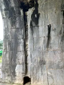 Đây là hình ảnh chụp cận cảnh một phần của một công trình kiến trúc bằng đá, có thể là một bức tường hoặc chân của một vòm cổng lớn như ở Thành nhà Hồ. Công trình được xây dựng từ những khối đá lớn, được đẽo gọt và xếp chồng lên nhau một cách chắc chắn. Bề mặt đá đã cũ, có nhiều vết nứt và bị phong hóa theo thời gian. Nhiều vệt nước màu đen và sẫm chảy dọc xuống, cùng với những mảng rêu xanh bám trên bề mặt, cho thấy sự tác động của thời tiết và độ ẩm. Ở phía dưới chân tường có một lỗ hổng nhỏ, tối. Phía sau bên trái có thể nhìn thấy một phần cây xanh và một công trình màu xanh lá cây nhưng không rõ chi tiết. Không có người hay hoạt động nào diễn ra trong ảnh.。（日本語訳）。これは石造りの建築物の一部を接写した画像で、胡朝の城のような大きなアーチ門の壁または土台である可能性があります。この建造物は、大きな石のブロックから造られており、それらは彫られ、しっかりと積み重ねられています。石の表面は古く、多くのひび割れがあり、時間とともに風化しています。多くの黒や濃い色の水の筋が縦に流れ落ち、表面に付着した緑の苔の斑点とともに、天候と湿度の影響を示しています。壁の付け根の下の方に、小さくて暗い穴があります。左奥には、緑の木の一部と緑色の建造物が見えますが、詳細は不明です。写真の中には人や活動は見られません。