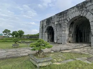 Hình ảnh cho thấy một công trình kiến trúc cổ bằng đá, đây là một phần của cổng Thành nhà Hồ ở Thanh Hóa. Cổng thành được xây dựng từ những khối đá lớn, có các lối vào hình vòm sâu. Bề mặt đá đã cũ và có nhiều vết bẩn do thời gian. Phía trước công trình là một bãi cỏ xanh và một con đường lát đá. Trên bãi cỏ có đặt hai chậu cây cảnh bonsai với chậu được trang trí hoa văn. Ở phía xa bên trái có cây cối và một tấm biển có chữ 'HỒ'. Bầu trời trong ảnh có nhiều mây trắng. Khung cảnh chung rất yên tĩnh, không có hoạt động nào của con người diễn ra.。（日本語訳）。写真は石造りの古い建築物を示しており、これはタインホア省にある胡朝の城の門の一部です。城門は大きな石のブロックから建てられており、深いアーチ型の入口があります。石の表面は古くなっており、時間の経過による多くの汚れがあります。建築物の前には緑の芝生と石畳の道があります。芝生の上には、模様で装飾された鉢に入った2つの盆栽が置かれています。左奥には木々と、「HỒ」という文字が書かれた看板があります。写真の空には白い雲がたくさんあります。全体的な光景はとても静かで、人の活動は見られません。