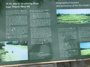 Đây là một tấm biển thông tin giới thiệu về "Vị trí địa lý và phong thủy của Thành Nhà Hồ", được đặt tại khu di tích. Tấm biển có nội dung bằng cả tiếng Việt và tiếng Anh, kèm theo các hình ảnh minh họa.。Nội dung chữ viết tiếng Việt được ghi lại như sau:。Tiêu đề: Vị trí địa lý và phong thủy của Thành Nhà Hồ。Nội dung chính:。"Di sản Thế Giới Thành Nhà Hồ ① nằm trên địa phận huyện Vĩnh Lộc. Vào thời Trần (1225-1400), vùng đất này có tên gọi là động An Tôn.。Động An Tôn là vùng đồng bằng rộng hàng nghìn héc-ta, được bao bọc bởi các dãy núi đá vôi phía Tây Bắc và hai con sông Mã, sông Bưởi phía Tây Nam và Đông Nam. Theo quan niệm phong thủy Phương Đông, đây là một vị thế và phong cảnh tuyệt đẹp, đắc địa cho việc chọn đất dựng đô.。Phía Nam Thành Nhà Hồ là nơi hội tụ của dòng sông Mã ② từ phía Tây chảy tới và sông Bưởi ③ từ phía Đông tụ về, được ví như “rồng chầu” và “rồng cuộn”. Hai con sông uốn quanh rồi tụ thủy ở phía Nam, tạo thành minh đường cho kinh đô.。Ở phía Nam Thành Nhà Hồ có dãy Đốn Sơn ④, hay núi Đún, được xem là tiền án.。Phía Bắc có dãy núi Thổ Tượng ⑤ (núi Voi) được xem là hậu chẩm.。Phía Đông có dãy núi Hắc Khuyển ⑥ (núi Chó đen) và phía Tây có dãy núi Ngọa Ngưu ⑦ (Trâu nằm), hay núi An Tôn được xem là “hổ phục tay ngai” án ngữ phía Tây ngôi thành.。Hồ Quý Ly chọn mảnh đất này thay thế cho Thăng Long bởi đây là vùng đất Thạch bàn - Long xà, nghĩa là thế đất như bàn đá, có rồng chầu sông Mã, rắn cuộn sông Bưởi. Đây là thế đất đẹp có vị trí bền vững, dài lâu.。Kinh thành mở ra bốn cổng: Đông, Tây, Nam và Bắc. Hướng chính của thành là hướng Nam. Đây là đặc điểm chung của các kinh thành phương Đông với quan niệm hoàng đế là bậc thiên tử ngoảnh mặt về phía Nam để cai trị thiên hạ. Theo quan niệm của người Việt, đây là hướng tốt lành, mang lại may mắn và sự yên vui.。Nối liền Cổng Nam là con đường Hoa Nhai (đường Hoàng Gia) ⑧ lát bằng đá phiến dài khoảng 2,5 km từ Cổng Nam tới tận đàn tế Nam Giao ⑨ ở Đốn Sơn."。Chú thích hình ảnh:。"Sông Bưởi là nhánh phụ của sông Mã có chiều dài khoảng 130 km. Cũng như sông Mã, sông Bưởi cũng có chức năng làm tuyến phòng ngự tự nhiên phía Đông Nam bảo vệ kinh thành."。Cảnh vật và hoạt động được mô tả trên biển:。- Địa điểm: Biển thông tin tại khu di tích Thành Nhà Hồ.。- Vật thể: Tấm biển thông tin, các hình ảnh chụp di tích và cảnh quan xung quanh.。- Hình ảnh 1: Chụp toàn cảnh di tích Thành Nhà Hồ từ trên cao, cho thấy nền móng của công trình kiến trúc cổ nằm giữa một vùng đồng bằng rộng lớn, bao quanh là ruộng lúa và các dãy núi.。- Hình ảnh 3: Chụp một đoạn sông (được chú thích là sông Bưởi) có màu nước xanh lục. Trên sông có một chiếc thuyền nhỏ đang hoạt động, có thể là thuyền đánh cá. Bờ sông là đất, có cây cối mọc ven bờ.。（日本語訳）。これは遺跡地区に設置された、「胡朝の城の地理的位置と風水」について紹介する案内板です。案内板にはベトナム語と英語の両方で内容が記載されており、説明のための写真が付いています。ベトナム語で書かれた内容は以下の通りです。タイトル：胡朝の城の地理的位置と風水。主な内容：。「世界遺産・胡朝の城①はヴィンロック県に位置しています。陳朝時代（1225年～1400年）、この地はアントン洞と呼ばれていました。アントン洞は数千ヘクタールに及ぶ広大な平野で、北西を石灰岩の山脈に、南西と南東をマー川とブオイ川の二つの川に囲まれています。東洋の風水の観念によれば、ここは都を建設する地を選ぶのに絶好の、非常に美しい地勢と風景を持つ場所です。胡朝の城の南側は、西から流れてくるマー川②と東から集まるブオイ川③が合流する場所であり、『龍の拝礼』と『龍の巻きつき』に例えられています。二つの川は蛇行しながら南で合流し、都のための明堂を形成しています。胡朝の城の南側にはドンソン山脈④、別名ドゥン山があり、前案と見なされています。北側にはトートゥオン山脈⑤（象山）があり、後枕と見なされています。東側にはハックエン山脈⑥（黒犬山）、西側にはゴアグウ山脈⑦（横たわる水牛）、別名アントン山があり、城の西側を守る『玉座の肘掛けに伏せる虎』と見なされています。胡季犛（ホー・クイ・リ）が昇龍（タンロン）に代わってこの地を選んだのは、ここが『石盤-龍蛇』の地、すなわち石の盤のような地勢で、マー川に拝礼する龍とブオイ川に巻きつく蛇がいる土地だからです。これは永続的で長期的な安定した位置を持つ美しい地勢です。都城には東、西、南、北の四つの門が開かれています。城の主要な方角は南向きです。これは、皇帝は天子であり、南を向いて天下を治めるという観念を持つ東洋の都城に共通する特徴です。ベトナム人の観念によれば、これは吉兆の方向であり、幸運と安らぎをもたらします。南門に繋がっているのは、南門からドンソンにある南郊壇⑨まで約2.5kmにわたって石板で舗装されたホアニャイ通り（王の道）⑧です。」。写真のキャプション：。「ブオイ川はマー川の支流で、長さは約130kmです。マー川と同様に、ブオイ川も都城を守る南東の自然な防衛線としての機能を持っています。」。案内板に描かれている風景と活動：。- 場所：胡朝の城遺跡地区にある案内板。- 物体：案内板、遺跡と周辺景観を撮影した写真。- 写真1：上空から撮影した胡朝の城遺跡の全景。広大な平野の真ん中に古代建築物の基礎部分があり、周囲を水田と山脈が囲んでいる様子がわかる。- 写真3：（ブオイ川と注釈がある）川の一部分を撮影したもの。川の水は緑色をしている。川の上には小さな船が一艘活動しており、漁船の可能性がある。川岸は土で、岸辺には木々が生えている。