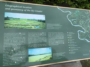 Đây là hình ảnh chụp cận cảnh một bảng thông tin giới thiệu về 'Vị trí địa lý và phong thủy của Thành nhà Hồ' (Geographical location and geomancy of the Ho Citadel). Bảng thông tin này có nền màu xanh lá cây, chữ màu trắng, được trình bày bằng cả tiếng Việt và tiếng Anh, đặt ở ngoài trời với cây cối xung quanh.。Nội dung trên bảng bao gồm:。1.  **Văn bản:** Các đoạn văn bản giải thích về địa thế của Thành nhà Hồ, các yếu tố phong thủy như sông và núi xung quanh. Phần chữ tiếng Việt có thể đọc được gồm:。*   Về Đàn tế Nam Giao: "Đàn tế Nam Giao được phát hiện ở chân núi Đún, cách thành nhà Hồ 2,5 km về phía Đông Nam. Đây từng là nơi hoàng đế làm lễ tế cầu quốc thái dân an hoặc đại xá thiên hạ."。*   Về Sông Mã: "Sông Mã ở thế kỷ 14-15 có tên là Lỗi Giang. Sông Mã là tuyến phòng ngự tự nhiên phía Tây Bắc bảo vệ kinh thành."。2.  **Bản đồ:** Một bản đồ chi tiết khu vực Huyện Vĩnh Lộc, thể hiện vị trí của Thành Nhà Hồ và các địa danh liên quan như Sông Mã, Sông Bưởi, Đốn Sơn (Núi Đún), Núi Thổ Tượng, Núi Hắc Khuyển, Núi Ngưu Ngọa, Đường Hoa Nhai, và Đàn tế Nam Giao. Các đơn vị hành chính như Xã Vĩnh Quang, Xã Vĩnh Yên, Xã Vĩnh Long, TT Vĩnh Lộc cũng được ghi trên bản đồ.。3.  **Hình ảnh:** Có hai bức ảnh màu. Một ảnh chụp toàn cảnh di tích Đàn tế Nam Giao, là một khu đất khảo cổ với các nền móng bằng đá trên bãi cỏ. Ảnh còn lại chụp Sông Mã với một vài nhà nổi hoặc bến thuyền nhỏ ven bờ.。4.  **Mã QR:** Ở góc dưới bên phải có một mã QR kèm chú thích: "Quét mã QR để khám phá thêm".。（日本語訳）。これは「胡朝の城の地理的位置と風水」（Geographical location and geomancy of the Ho Citadel）を紹介する案内板のクローズアップ写真です。この案内板は緑色の背景に白文字で、ベトナム語と英語の両方で書かれており、周囲に木々がある屋外に設置されています。案内板の内容は以下の通りです。1.  **テキスト：**胡朝の城の地勢、周囲の川や山などの風水的要素について説明する文章。読み取れるベトナム語の部分は以下の通りです。*   南郊壇について：「南郊壇は、胡朝の城から南東に2.5km離れたドゥン山の麓で発見されました。ここはかつて皇帝が国泰民安を祈願したり、天下に大赦を行ったりするための祭祀を行った場所でした。」。*   マー川について：「14世紀から15世紀にかけて、マー川はロイ川という名前でした。マー川は都を保護する北西の自然な防衛線です。」。2.  **地図：**ヴィンロック県の詳細な地図で、胡朝の城の位置と、マー川、ブオイ川、ドンソン（ドゥン山）、トー・トゥオン山、ハク・クエン山、グウ・ゴア山、ホア・ニャイ通り、南郊壇などの関連する地名が示されています。ヴィン・クアン社、ヴィン・イエン社、ヴィン・ロン社、ヴィンロック町などの行政単位も地図に記載されています。3.  **写真：**カラー写真が2枚あります。1枚は南郊壇遺跡の全景を撮影したもので、芝生の上に石の基礎がある考古学的な敷地です。4.  **QRコード：**右下隅に「QRコードをスキャンしてさらに詳しく」というキャプション付きのQRコードがあります。