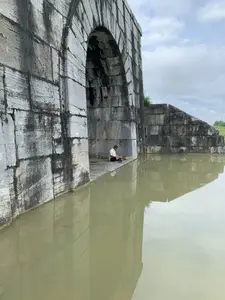 Bối cảnh là một công trình kiến trúc cổ bằng đá, cụ thể là một cổng thành của Thành nhà Hồ. Cổng thành được xây dựng từ những khối đá lớn, vững chãi, đã nhuốm màu thời gian với những vết ố đen. Bên cạnh cổng thành là một hào nước, mặt nước tĩnh lặng và có màu đục, phản chiếu hình ảnh của cổng thành và bầu trời nhiều mây. Bên trong một mái vòm của cổng thành, một người đàn ông lớn tuổi đang ngồi xếp bằng trên nền đá, ngay sát mép nước. Ông mặc áo sơ mi sáng màu và quần tối màu. Bên cạnh ông có một chiếc nón. Người đàn ông dường như đang nghỉ ngơi hoặc có thể đang câu cá. Không có chữ viết hay văn bản nào có thể nhận dạng được trong hình ảnh.。（日本語訳）。背景は石造りの古い建築物で、具体的には胡朝の城の城門です。城門は大きく頑丈な石のブロックで建てられており、黒いシミがついて時の色に染まっています。城門の隣には堀があり、水面は静かで濁った色をしており、城門と曇り空の姿を映しています。城門のアーチの内側で、一人の年配の男性が水際のすぐそばの石の床にあぐらをかいて座っています。彼は明るい色のシャツと暗い色のズボンを履いています。彼の隣には笠が一つあります。その男性は休んでいるか、あるいは釣りをしているようです。画像の中には、認識できる文字や文章はありません。