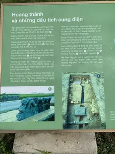 Đây là hình ảnh chụp một bảng thông tin tại khu di tích lịch sử Thành Nhà Hồ. Bảng thông tin có nền màu xanh lá cây, chữ màu trắng, và khung kim loại, được đặt ngoài trời trên một bệ bê tông. Bảng cung cấp thông tin về các di tích của hoàng thành.。**Nội dung chữ viết được ghi lại từ bảng thông tin:**。**Hoàng thành và những dấu tích cung điện**。Bạn đang nhìn về hướng Bắc của Thành Nhà Hồ. Khi thời tiết tốt, từ đây bạn có thể nhìn thấy Cổng Bắc và đôi rồng đá thềm bậc ①.。Hoàng thành, hay còn gọi Thành nội, là nơi thiết triều và sinh hoạt của triều đình.。Vòng ngoài Thành nội được bao bọc bằng hệ thống Hào thành ② và La thành ③, trải rộng trên diện tích 5.480 héc ta.。Chính giữa Hoàng thành từng là chính điện (nơi thiết triều), mà dấu tích khảo cổ hiện còn thấy ở Nền Vua ④ và đôi rồng đá thềm bậc được cho là của chính điện ⑤.。Tương tự kinh đô Thăng Long từ nhà Lý (1010-1224) đến nhà Trần (1225-1400), các kiến trúc cung điện chính được bố trí cân đối ở hai bên chính điện.。Phía trước chính điện có Đông Thái Miếu và Tây Thái Miếu. Đông Thái Miếu là nơi thờ tổ tiên họ Hồ, còn Tây Thái Miếu thờ họ ngoại (Trần Minh Tông và Trần Nghệ Tông).。Phía sau chính điện, bên hữu (bên phải) có cung Nhân Thọ là nơi ở của hoàng đế, bên tả (bên trái) có điện Hoàng Nguyên là nơi hoàng đế làm việc hàng ngày.。Trong Thành nội còn có nhiều kiến trúc khác như Đông Cung (nơi ở của thái tử) và cung Phù Cực (nơi ở của hoàng hậu).。Trong thành đường đi lối lại đều được lát đá. Một số hồ được đào để tạo phong cảnh như hồ Dục Thúy ⑥, hồ Bơi Chải ⑦, hồ Dục Tượng ⑧ và hồ Bán Nguyệt ⑨.。Hồ Bán Nguyệt có ý nghĩa hài hòa âm dương cho tòa thành, nằm cạnh điện Hoàng Nguyên nơi hoàng đế làm việc hàng ngày.。**Mô tả các hình ảnh trên bảng:**。1.  **Hình ảnh ①:** Chụp một đôi rồng đá điêu khắc ở hai bên thềm bậc. Phía sau đôi rồng là một con đường lát gạch và xa hơn là một cánh đồng lúa xanh.。2.  **Hình ảnh ④:** Chụp một hố khai quật khảo cổ học nhìn từ trên xuống. Bên trong hố lộ ra các dấu tích nền móng, tường và các cấu trúc bằng đá của một công trình kiến trúc cổ.。（日本語訳）。これは胡朝の城歴史遺跡地区にある案内板を撮影した写真です。案内板は緑色の背景に白い文字、金属のフレームがあり、コンクリートの台座の上に屋外に設置されています。この板は皇城の遺跡に関する情報を提供しています。**案内板から書き起こした文字の内容：**。**皇城と宮殿の痕跡**。あなたは胡朝の城の北側を見ています。天気が良い時、ここから北門と階段の石の龍のペア①を見ることができます。皇城、または城内とも呼ばれ、朝廷が政務を行い、生活する場所でした。城内の外周は、城壕②と羅城③のシステムによって囲まれており、5,480ヘクタールの面積に広がっています。皇城の中央にはかつて正殿（政務を行う場所）があり、その考古学的痕跡は現在、皇帝の nền（Nền Vua）④と、正殿のものとされる階段の石の龍のペア⑤に見られます。李朝（1010-1224）から陳朝（1225-1400）までの昇龍京城と同様に、主要な宮殿建築は正殿の両側に均等に配置されていました。正殿の前には東太廟と西太廟がありました。東太廟は胡氏の祖先を祀る場所であり、一方、西太廟は母方の血縁（陳明宗と陳芸宗）を祀っていました。正殿の後ろ、右側（右）には皇帝の住居である仁寿宮があり、左側（左）には皇帝が日常の業務を行う場所である皇元殿がありました。城内には他にも、東宮（皇太子の住居）や浮極宮（皇后の住居）など多くの建築物がありました。城内の通路はすべて石で舗装されていました。景観を作るために、浴翠池⑥、ボイチャイ池⑦、浴象池⑧、半月池⑨などのいくつかの池が掘られました。半月池は城の陰陽の調和を意味し、皇帝が日常の業務を行う皇元殿の隣にありました。**板上の写真の説明：**。1.  **写真①：** 階段の両側にある一対の石の龍の彫刻を撮影したもの。龍のペアの後ろにはレンガ敷きの道があり、さらにその先には緑の田んぼが広がっています。2.  **写真④：** 考古学的な発掘坑を上から撮影したもの。坑内には、古代の建築物の基礎、壁、石造りの構造物の痕跡が現れています。