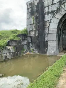 Bức ảnh chụp một công trình kiến trúc cổ bằng đá lớn, có thể là một phần của một thành cổ hoặc một cây cầu. Cấu trúc được xây dựng từ những khối đá hình chữ nhật lớn, đã bị phong hóa theo thời gian với những vệt ố đen và cây dại mọc trong các kẽ hở. Ở bên phải, có một lối vào hình vòm dẫn vào một đường hầm tối. Phía trước công trình là một con hào hoặc kênh nước tù đọng, có màu nâu đục. Bờ đất ven con hào và ngọn đồi nhỏ ở phía sau được bao phủ bởi cỏ xanh. Bầu trời u ám và nhiều mây. Không có bất kỳ văn bản hay hoạt động nào của con người được ghi nhận trong ảnh.。（日本語訳）。写真は、古代の城壁または橋の一部と思われる、大きな石でできた古い建築物を写しています。この建造物は、大きな長方形の石のブロックで造られており、時間とともに風化し、黒い染みができ、隙間には雑草が生えています。右側には、暗いトンネルへと続くアーチ型の入り口があります。建造物の前には、濁った茶色のよどんだ水がある堀または水路があります。堀沿いの土手と後方の小さな丘は緑の草で覆われています。空はどんよりとしており、雲が多いです。写真には、いかなる文字や人間の活動も記録されていません。