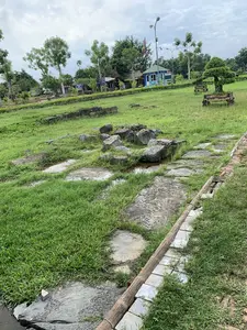 Đây là một khu di tích lịch sử hoặc công viên khảo cổ. Bức ảnh chụp một bãi cỏ rộng với những phiến đá cổ và tàn tích bằng đá. Một con đường lát đá, một phần bị ngập nước và cỏ mọc xen kẽ, chạy qua khu vực. Ở hậu cảnh, có một ki-ốt bán vé màu xanh lam với dòng chữ có thể đọc được là 'BÁN VÉ THAM QUAN KHU DU LỊCH' và 'HỒ CÁ SẤU KHU DU LỊCH'. Gần đó có một cột cờ treo lá cờ Việt Nam. Xung quanh có nhiều cây xanh, cột đèn đường, một cây cảnh bonsai trong chậu trang trí và những chiếc ghế đá có hoa văn. Không có người hoặc hoạt động cụ thể nào đang diễn ra, khung cảnh tĩnh lặng, cho thấy đây là một địa điểm tham quan.。（日本語訳）。これは歴史的遺跡または考古学公園です。石畳の道が、一部水に浸かり、草が点在しながら、そのエリアを横切っています。背景には、「BÁN VÉ THAM QUAN KHU DU LỊCH」（観光地入場券販売）と「HỒ CÁ SẤU KHU DU LỊCH」（ワニ池観光地）と読める文字が書かれた青いチケット売り場があります。その近くには、ベトナム国旗を掲げた旗竿があります。周囲には多くの緑の木々、街灯、装飾的な鉢に入った盆栽、そして模様のある石のベンチがあります。人や特定の活動は見られず、静かな光景は、ここが観光地であることを示唆しています。
