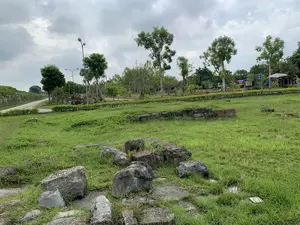 Quang cảnh ngoài trời tại một khu di tích lịch sử, có khả năng là khu vực Thành nhà Hồ. Bầu trời nhiều mây. Tiền cảnh là một bãi cỏ xanh rộng với nhiều tảng đá lớn, có thể là tàn tích của một công trình cổ, nằm rải rác. Một phần móng tường thấp bằng đá cũng hiện diện. Ở phía sau, có một con đường đất chạy dọc theo một bức tường thành bằng đá cao hơn. Khu vực này được bài trí như công viên với cây xanh, hàng rào cây được cắt tỉa, cột đèn đường, và một chiếc xe kéo gỗ trang trí. Có một lá cờ Việt Nam cắm giữa các lùm cây. Có các biển báo trong khu vực. Một biển báo ghi 'LỐI VÀO BÃI GỬI XE'. Một biển chỉ dẫn khác liệt kê các địa điểm: 'Đền thờ Bình Khương', 'Giếng Vua', 'Cổng Đông Môn', 'Cổng Tây Môn', 'Cổng Nam Môn', 'Nhà trưng bày', 'WC', và có biểu tượng bãi đỗ xe (P). Khung cảnh yên tĩnh, không có hoạt động nào đáng kể.。（日本語訳）。ある史跡の屋外風景、胡朝の城の区域である可能性が高い。空は曇りが多い。前景には広大な緑の芝生が広がり、古代の建造物の遺跡と思われる大きな石が点在している。石でできた低い壁の基礎部分も見られる。後方には、より高い石の城壁に沿って未舗装の道が続いている。この区域は、緑の木々、刈り込まれた生け垣、街灯、そして装飾用の木製荷車があり、公園のように整備されている。茂みの中にベトナムの国旗が一本立てられている。区域内には標識がある。ある標識には「駐車場入口」と書かれている。別の案内標識には、「ビンクオン祠」、「王の井戸」、「東門」、「西門」、「南門」、「展示館」、「WC」といった場所がリストアップされており、駐車場（P）のシンボルも表示されている。風景は静かで、特筆すべき活動は見られない。