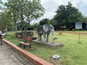 Trong ảnh có một tấm biển hiệu lớn màu xanh của nhà hàng với nội dung: 'NHÀ HÀNG TÂY ĐÔ THÀNH NHÀ HỒ. CHUYÊN CÁC MÓN ĂN ĐẶC SẢN VĨNH LỘC. NHẬN ĐẶT TIỆC LIÊN HOAN, SINH NHẬT, HỘI NGHỊ. CƠM ĐẶT - CƠM BÌNH DÂN. PHỤC VỤ TOUR DU LỊCH. ĐC: CỔNG NAM THÀNH NHÀ HỒ - XUÂN GIAI, VĨNH TIẾN, VĨNH LỘC, THANH HÓA. ĐT: 0969 401 151 / 0919 911 656'.。Bối cảnh là một không gian ngoài trời, có thể là công viên hoặc khuôn viên của một khu di tích lịch sử. Nổi bật ở giữa là một bức tượng voi lớn bằng xi măng màu xám, có ngà, đang đứng trên bệ. Phần vòi của tượng voi bị hư hỏng, để lộ cốt thép bên trong. Xung quanh là bãi cỏ xanh, cây cối, một băng ghế, và một số khối đá lớn được trưng bày. Có một lối đi bộ lát đá và một người đàn ông đang đứng ở bên trái, dường như đang tham quan.。（日本語訳）。写真には、レストランの大きな青い看板があり、その内容は「レストラン・タイドー・胡朝の城。ヴィンロック特産料理専門店。宴会、誕生日、会議の予約受付。予約弁当 - 大衆食堂。観光ツアーへのサービス提供。住所：タインホア省ヴィンロック県ヴィンティエン社スアンザイ村 - 胡朝の城南門。電話番号：0969 401 151 / 0919 911 656」です。背景は屋外の空間で、公園か史跡の敷地内である可能性があります。中央で目立っているのは、牙があり、台座の上に立っている、灰色のセメントでできた大きな象の像です。象の像の鼻の部分は損傷しており、中の鉄筋が露出しています。周りには緑の芝生、木々、一つのベンチ、そして展示されているいくつかの大きな石の塊があります。石畳の遊歩道があり、左側には一人の男性が立っていて、見学しているようです。