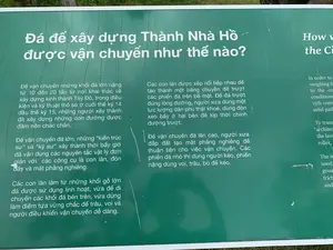 Hình ảnh chụp cận cảnh một bảng thông tin màu xanh lá cây với chữ trắng tại khu di tích Thành Nhà Hồ. Bảng thông tin này giải thích về các phương pháp được sử dụng để vận chuyển đá xây dựng thành. Toàn bộ nội dung văn bản tiếng Việt trên bảng được ghi lại như sau:。Đá để xây dựng Thành Nhà Hồ được vận chuyển như thế nào。Để vận chuyển những khối đá lớn nặng từ 10 đến 20 tấn từ nơi khai thác về xây dựng kinh thành Tây Đô, trong điều kiện và kỹ thuật thô sơ ở cuối thế kỷ 14 đầu thế kỷ 15, những người xây thành đã xây dựng những con đường được đầm nền chắc chắn.。Để vận chuyển đá lớn, những “kiến trúc sư” và “kỹ sư” xây thành thời bấy giờ đã vận dụng các nguyên tắc vật lý đơn giản với các công cụ là con lăn, đòn bẩy và mặt phẳng nghiêng.。Các con lăn làm từ những khối gỗ lớn đã được sử dụng linh hoạt, vừa để di chuyển các khối đá bên trên, vừa dùng làm điểm tựa vững chắc để trâu, voi và người điều khiển vận chuyển dễ dàng.。Các con lăn được xếp nối tiếp nhau để tạo thành một băng chuyền để trượt các phiến đá trên bề mặt. Để đá trượt đúng lòng đường, người xưa dùng một lực lượng dân phu thật khoẻ, dùng đòn xeo bẩy ở hai bên để kịp thời chỉnh đường trượt.。Để vận chuyển đá lên cao, người xưa đắp đất tạo mặt phẳng nghiêng để thuận tiện cho việc vận chuyển. Các phiến đá nhỏ thì dùng người kéo, phiến nặng dùng voi, trâu, bò để kéo.。（日本語訳）。胡朝の城跡にある、白い文字が書かれた緑色の案内板のクローズアップ写真。この案内板は、城の建設に使われた石を運搬するために使用された方法について説明しています。案内板に書かれているベトナム語のテキスト全文は以下の通りです。胡朝の城を建設するための石はどのように運ばれたのか。14世紀末から15世紀初頭の原始的な条件と技術の中で、採石場から10トンから20トンの重さの巨大な石塊を西都（タイドー）の都城建設地まで運ぶために、城の建設者たちは固く突き固められた道を建設しました。大きな石を運ぶために、当時の城の建設における「建築家」や「技術者」たちは、コロ、てこ、斜面といった道具を用いて、単純な物理法則を応用しました。大きな木材から作られたコロは柔軟に使用され、上の石塊を移動させるためだけでなく、水牛、象、そして操作する人々が容易に運搬できるよう、しっかりとした支点としても機能しました。コロは連続して並べられ、その表面で石板を滑らせるためのコンベアを形成しました。石が道筋から外れずに滑るように、昔の人々は非常に屈強な労働者を使い、両側からてこ棒でこじり、滑る方向を適時修正しました。石を高い場所へ運ぶために、昔の人々は土を盛って斜面を作り、運搬を容易にしました。小さな石板は人力で引き、重い石板は象、水牛、牛を使って引きました。