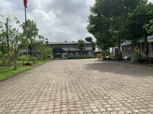 Bức ảnh chụp một khoảng sân rộng phía trước một tòa nhà, đây là Trung tâm Trưng bày Di sản Văn hóa Thế giới Thành Nhà Hồ. Trên vòm cửa lối vào chính của tòa nhà có dòng chữ: "TRUNG TÂM TRƯNG BÀY DI SẢN VĂN HÓA THẾ GIỚI THÀNH NHÀ HỒ" và bên dưới là dòng chữ tiếng Anh "Exhibition of the Citadel of the Ho Dynasty". Sân được lát bằng gạch khối hình bát giác. Bên trái sân có một cột cờ cao đang treo một lá cờ đỏ. Tòa nhà trưng bày là một công trình một tầng, trên tường phía ngoài có gắn các tấm pa-nô lớn in hình ảnh các hiện vật. Xung quanh sân được trồng nhiều cây xanh. Bầu trời trong ảnh có nhiều mây. Khung cảnh vắng lặng, không có người hay hoạt động nào đang diễn ra.。（日本語訳）。写真は、ある建物の前にある広い中庭を写しており、これは胡朝の城塞世界文化遺産展示センターです。建物の正面玄関のアーチの上には、「TRUNG TÂM TRƯNG BÀY DI SẢN VĂN HÓA THẾ GIỚI THÀNH NHÀ HỒ」という文字があり、その下には英語で「Exhibition of the Citadel of the Ho Dynasty」と書かれています。中庭は八角形のブロックタイルで舗装されています。中庭の左側には、赤い旗が掲げられた高い旗竿があります。展示棟は1階建ての建物で、外壁には遺物の写真が印刷された大きなパネルが取り付けられています。中庭の周りには多くの緑の木が植えられています。写真の空は雲が多いです。風景は静かで、人や活動は見られません。