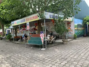 Đây là một quán giải khát hoặc trạm dừng chân ven đường. Phía trước quán có mái hiên, dưới mái hiên có vài người đang ngồi nghỉ trên ghế nhựa và ghế dài. Một chiếc xe máy đang đỗ gần đó. Sân được lát bằng gạch block hình bát giác. Quán được trang trí bằng những chiếc đèn lồng đỏ. Trên tường bên hông có một bức tranh vẽ di tích Thành nhà Hồ.。Các dòng chữ có thể đọc được trên các biển hiệu bao gồm:。- Biển hiệu chính dưới mái hiên: "ĐẶC SẢN: BÁNH RAU MÁ - BÁNH RĂNG BỪA - CHÈ LAM - KẸO LẠC PHÚ QUANG".。- Biển hiệu phía trên: "OCOP... NAM" và "ĐẶC SẢN KEM CHUA".。- Biển hiệu nhỏ màu đỏ: "ĐẶC SẢN NEM CHUA".。- Biển hiệu đứng: "KEM SỮA CHUA CÀ PHÊ NƯỚC NGỌT Các loại" và một biển hiệu khác của "Aice KEM THƯƠNG HIỆU SINGAPORE".。- Trên các ô che nắng có logo "Coca-Cola".。（日本語訳）。これは道端の軽食店または休憩所です。店の前には軒があり、その下では数人がプラスチックの椅子や長椅子に座って休んでいます。近くにバイクが1台停まっています。庭は八角形のブロックタイルで舗装されています。店は赤い提灯で飾られています。側面の壁には、胡朝の城跡を描いた絵があります。看板で読み取れる文字は以下の通りです：。- 軒下のメイン看板：「特産品：バイン・ラウマー - バイン・ランブア - チェー・ラム - フークアンのピーナッツ菓子」。- 上部の看板：「OCOP... NAM」と「特産品 サワークリーム」。- 小さな赤い看板：「特産品 ネムチュア」。- 立て看板：「アイスクリーム、ヨーグルト、コーヒー、ソフトドリンク 各種」と、もう一つの「Aice シンガポールブランドのアイスクリーム」の看板。- 日よけのパラソルには「コカ・コーラ」のロゴがあります。