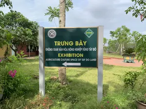 Hình ảnh chụp một tấm biển chỉ dẫn ngoài trời tại một khu vực có vẻ là công viên hoặc khu di tích văn hóa. Biển báo có nền màu xanh lá cây, được dựng trên hai cột màu xám.。Nội dung trên biển báo được ghi lại như sau:。Phần chữ chính bằng tiếng Việt, màu vàng: 'TRƯNG BÀY - KHÔNG GIAN VĂN HÓA NÔNG NGHIỆP VÙNG TÂY ĐÔ'.。Phần dịch tiếng Anh bên dưới, màu trắng: 'EXHIBITION - AGRICULTURE CULTURE SPACE OF TAY DO LAND'.。Có một mũi tên lớn màu trắng chỉ hướng sang trái.。Ở góc trên bên trái là logo Di sản Thế giới của UNESCO với dòng chữ 'DI SẢN THẾ GIỚI - WORLD HERITAGE - PATRIMOINE MONDIAL'.。Ở góc trên bên phải là logo của Thành nhà Hồ với dòng chữ 'THE HO CITADEL - THÀNH NHÀ HỒ'.。Bối cảnh xung quanh biển báo là một không gian xanh mát với bãi cỏ, nhiều cây cối và các bụi hoa (có hoa giấy màu hồng ở phía trước). Phía sau, có một con đường lát gạch màu đỏ, một chậu cây cảnh lớn và có thể thấy lờ mờ các bức tượng hình con trâu, phù hợp với chủ đề 'Không gian văn hóa nông nghiệp'.。（日本語訳）。公園または文化遺跡と思われる屋外エリアにある案内看板を撮影した写真。看板は緑色の背景で、灰色の2本の柱の上に設置されています。看板に記載されている内容は以下の通りです。ベトナム語の主要な黄色い文字：「展示 - 西都（タイドー）地域農業文化空間」。その下の白色の英語訳：「EXHIBITION - AGRICULTURE CULTURE SPACE OF TAY DO LAND」。左を指す大きな白い矢印があります。左上隅には、「DI SẢN THẾ GIỚI - WORLD HERITAGE - PATRIMOINE MONDIAL」という文字が入ったUNESCOの世界遺産ロゴがあります。右上隅には、「THE HO CITADEL - THÀNH NHÀ HỒ」という文字が入った胡朝の城塞のロゴがあります。看板の周りの背景は、芝生、多くの木々、花の茂みがある緑豊かな空間です（手前にはピンク色のブーゲンビリアがあります）。後方には、赤いレンガで舗装された道、大きな鉢植えの観葉植物があり、「農業文化空間」というテーマにふさわしい水牛の形をした像がぼんやりと見えます。