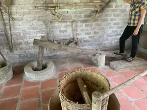Bối cảnh là một không gian trong nhà với tường gạch mộc và nền lát gạch đỏ, có vẻ như là một khu trưng bày hoặc bảo tàng về nông cụ truyền thống. Các vật thể chính trong ảnh bao gồm một bộ cối giã gạo bằng đá và gỗ kiểu cũ. Bộ cối này có một đòn bẩy bằng gỗ dài, một đầu là chày gỗ được đặt trong một cối đá. Ở phía trước, có một chiếc rổ lớn đan bằng tre. Treo trên tường là các nông cụ cũ khác như ách trâu bò và liềm. Một người mặc áo sơ mi kẻ sọc vàng xanh và quần đen đang đứng ở phía bên phải, dường như đang quan sát hoặc trình diễn cách sử dụng các dụng cụ. Trên một nhãn giấy dán trên dụng cụ trong rổ tre có ghi chữ: 'ÔNG KÉO KHÔNG XOAY' và 'CỐI GIÃ GẠO'. Hoạt động đang diễn ra là trưng bày các nông cụ truyền thống của Việt Nam.。（日本語訳）。背景は、むき出しのレンガ壁と赤レンガの床を持つ屋内空間で、伝統的な農具の展示エリアまたは博物館のようです。写真の主な物体には、石と木でできた旧式の米つき臼一式が含まれています。この臼一式には、長い木製のてこがあり、その一端には石臼の中に置かれた木製の杵があります。手前には、竹で編まれた大きなかごがあります。壁には、牛や水牛のくびきや鎌など、他の古い農具が掛けられています。黄色と緑の縞模様のシャツと黒いズボンを履いた人が右側に立っており、道具の使い方を観察または実演しているようです。竹かごの中の道具に貼られた紙のラベルには、「ÔNG KÉO KHÔNG XOAY」と「CỐI GIÃ GẠO」という文字が書かれています。行われている活動は、ベトナムの伝統的な農具の展示です。