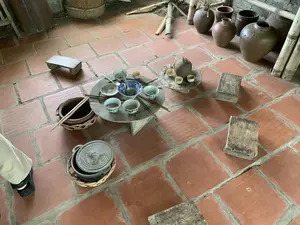 Bối cảnh là một không gian trong nhà với sàn được lát bằng gạch tàu màu đỏ, tái hiện lại một góc sinh hoạt của một gia đình Việt Nam xưa. Ở trung tâm, một mâm cơm được bày trên một chiếc bàn tròn thấp, có thể là một cái cối xay cũ. Trên mâm có nhiều bát sứ, một số có hoa văn xanh trắng, cùng với đũa và thìa. Gần đó là một khay trà riêng với một ấm trà và các chén nhỏ bằng gốm. Xung quanh trên sàn nhà có rải rác vài chiếc ghế đẩu gỗ thấp. Ngoài ra còn có một chiếc nồi gang có nắp đậy và một chiếc nồi đất, cả hai đều được đặt trong giỏ đan bằng tre. Ở phía sau là nhiều chum, vại gốm và các cây tre dựng dựa vào tường. Cảnh vật được sắp đặt, mô phỏng một bữa ăn hoặc một buổi quây quần uống trà theo lối sống truyền thống. Không có chữ viết nào có thể đọc được.。（日本語訳）。背景は、赤いテラコッタタイルが敷かれた屋内の空間で、昔のベトナムの家庭の生活の一角を再現している。中央には、低い丸テーブル、おそらく古い石臼の上に食事のお膳が並べられている。お膳の上には多くの磁器の碗があり、そのうちのいくつかは青と白の模様があり、箸とスプーンも添えられている。その近くには、急須と陶器の小さな湯呑みが乗った別のお茶の盆がある。床の周りには、いくつかの低い木製のスツールが点在している。その他に、蓋付きの鋳鉄鍋と土鍋があり、どちらも竹で編んだ籠の中に入れられている。後ろには、多くの陶器の壺や甕、そして壁に立てかけられた竹の棒がある。この光景は、伝統的な生活様式に沿った食事、または集まってお茶を飲む会を模して配置されている。読み取れる文字はない。