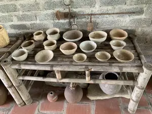 Bối cảnh là một không gian trưng bày trong nhà, có thể là một phần của bảo tàng hoặc nhà cổ, với bức tường xây bằng gạch đá xỉ và nền lát gạch đỏ. Trên một chiếc chõng tre (kệ tre) cũ, có trưng bày một bộ sưu tập gồm nhiều bát đĩa gốm sứ với các kích cỡ khác nhau. Các vật dụng này trông cũ kỹ, mộc mạc, một số đã bị sứt mẻ. Treo trên tường phía sau là một chiếc cân đòn gánh (cân ta) kiểu cũ, đã bị hoen gỉ. Bên dưới chõng tre, có đặt vài chiếc vò, chum sành. Một chiếc vò nhỏ có dán nhãn ghi chữ: "Vò đựng hạt giống".。（日本語訳）。背景は、博物館か古民家の一部であるかもしれない屋内の展示空間で、壁はスラグ煉瓦で造られ、床には赤レンガが敷かれている。古い竹製の寝台（竹製の棚）の上には、様々なサイズの陶磁器の碗や皿からなるコレクションが展示されている。これらの品々は古びて素朴に見え、いくつかは欠けている。後ろの壁には、錆びついた旧式の天秤棒（伝統的な秤）が一つ掛かっている。小さな壺の一つには「種入れの壺」と書かれたラベルが貼られている。