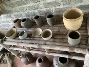 Bức ảnh chụp một bộ sưu tập đồ gốm, sành sứ cũ được trưng bày trên một chiếc kệ tre hai tầng. Các đồ vật này bao gồm nhiều loại với kích thước và hình dáng khác nhau như chum, vại, hũ, và bát. Chúng có màu sắc mộc mạc như nâu, xám, và vàng đất, trông cũ và mang dấu ấn thời gian. Chiếc kệ tre được đặt sát một bức tường xây bằng gạch xỉ chưa trát, và sàn nhà được lát gạch đỏ. Không có người hay hoạt động cụ thể nào diễn ra, đây là một cảnh tĩnh trưng bày các đồ vật thủ công truyền thống. Không có thông tin dạng chữ viết nào trong ảnh.。（日本語訳）。写真は、二段の竹製の棚の上に展示されている古い陶器や磁器のコレクションを写しています。これらの品物には、甕、壺、瓶、鉢など、さまざまなサイズや形のものが含まれています。それらは茶色、灰色、土黄色といった素朴な色合いで、古びて見え、時の痕跡を帯びています。竹製の棚は、漆喰が塗られていない燃え殻レンガで造られた壁際に置かれており、床には赤いタイルが敷かれています。人や特定の活動は見られず、伝統的な工芸品を展示している静的な光景です。写真には文字情報はありません。