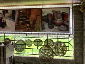 写真はセメントレンガの壁のある部屋の中から、大きな窓枠を通して外を眺めるように撮られています。窓の外には、ベトナムの農村地域に特有の、青々とした水田の風景が広がっています。部屋の中では、装飾品や写真がすべて伝統文化の特色を色濃く帯びています。窓の前には、竹やニッパヤシで編まれた円形の多くの道具が吊るされており、それらは魚を捕るための「lờ」や「đó」（魚捕りかご）、あるいは装飾用に使われる「rổ rá」（ざるやかご）かもしれません。窓の上には、額装された3枚組の大きな写真があり、手工芸の活動を描写しています。左の写真は藁ぼうきを作っているおばあさんを写しています。真ん中の写真はかまどと藁製品のある生活の光景です。右の写真は「chum」（水甕）、「vại」（甕）、「nồi đất」（土鍋）のような多くの陶磁器製品を展示しています。写真の中には読み取れる文字はありません。空間全体が、伝統工芸村にある展示室、あるいは文化体験スペースのイメージを想起させます