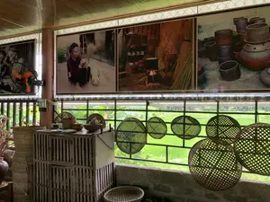 背景は、農村部にある手工芸品を展示する家または工房の内部空間です。大きな窓枠からは、青々とした水田が見えます。内部には、竹や籐でできた多くの品物が展示されており、さまざまなサイズやデザインのかご、ざる、フードカバー、編みかごが含まれています。いくつかの円形のフードカバーが装飾として吊るされています。その他にも、古い木製の棚、壁掛け扇風機、そしてテト（旧正月）を飾るための桃の造花の枝があります。壁には、大判の写真が一列に掛けられています。これらの写真は、伝統的な工芸村の生活と仕事を描写しています。ある写真は、座って手作りのほうきを作っている老婆を写し、別の写真は、かまどのある家の中での生活風景を写し、そしてもう一枚の写真は、甕（かめ）や壺（つぼ）のような多くの陶磁器製品を展示しています。全体の光景は、ベトナムの田舎の村の文化が色濃く感じられる空間を思い起こさせます