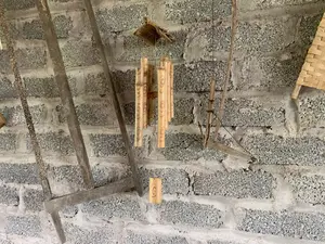 Hình ảnh cho thấy một bức tường gạch xỉ (gạch bê tông cốt liệu) thô, chưa được trát vữa. Trên bức tường này treo nhiều đồ vật trang trí mang tính mộc mạc. Ở vị trí trung tâm là một bộ chuông gió làm bằng tre, với các ống tre được khắc họa tiết xoắn ốc. Bên trái là một vật dụng bằng gỗ cũ, trông giống một phần của nông cụ. Bên phải cũng có một vật trang trí bằng gỗ, có hình dáng giống một công cụ nông nghiệp thu nhỏ. Góc trên bên phải có một phần của một vật đan bằng tre. Không có hoạt động nào diễn ra; các đồ vật này được treo cố định để trang trí.。（日本語訳）。写真には、漆喰が塗られていない粗いスラグレンガ（骨材コンクリートブロック）の壁が写っている。この壁には、多くの素朴な装飾品が掛けられている。中央には竹製の風鈴があり、その竹筒には螺旋模様が彫られている。右側にもまた、小型の農具のような形をした木製の装飾品がある。右上隅には、竹で編まれた物の一部がある。何の動きもなく、これらの物は装飾のために固定して掛けられている。