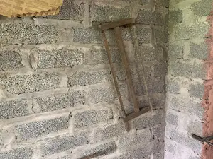 Bức ảnh chụp cận cảnh một góc tường được xây bằng gạch xỉ (gạch bê tông) chưa trát vữa. Dựa vào góc tường là một chiếc cưa khung bằng gỗ đã cũ, có phần lưỡi cưa bị gỉ sét. Ở góc trên bên trái của bức ảnh, có thể thấy một phần của một vật dụng được đan bằng tre, trông giống như một cái nia hoặc cái mẹt. Không có người hay hoạt động nào được ghi lại trong ảnh. Không có bất kỳ chữ viết nào.。（日本語訳）。漆喰が塗られていないスラグレンガ（コンクリートブロック）で造られた壁の角をクローズアップで撮影した写真。壁の角には、刃の部分が錆びた古い木製のフレームソーが立てかけられている。写真には人や活動は記録されていない。いかなる文字もない。