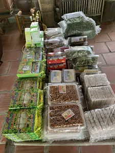 Hình ảnh chụp một quầy hàng trưng bày và bán nhiều loại đặc sản địa phương, được đặt trên một chiếc bàn hoặc chõng tre trên nền gạch đỏ. Các sản phẩm được đóng gói cẩn thận, sẵn sàng để bán.。Các vật phẩm được trưng bày bao gồm:。- Các hộp "Bánh Rau Má Đặc Sản Sầm Sơn" của "Cơ sở sản xuất bánh kẹo Kim Thành", một số hộp có nhãn "Hàng VIP" và "Xuất Khẩu".。- Các hộp "Chè Lam Phủ Quảng" của thương hiệu "Cương Xuân", có logo "OCOP" (Mỗi xã một sản phẩm).。- Các khay kẹo lạc (kẹo đậu phộng) được đóng gói trong bao bì nhựa trong suốt, có nhãn hiệu "Lạc Thảo" và cũng là sản phẩm OCOP.。- Nhiều loại thảo mộc sấy khô được đóng trong túi ni lông, bao gồm "Mướp đắng rừng", "Sen khô", và các túi lớn chứa lá cây khô.。- Các loại bánh kẹo khác như "Bánh đa Cương Xuân" và các loại bánh hình chữ nhật được bọc trong túi ni lông.。Trong hậu cảnh, có một chiếc chum sành lớn màu nâu, một chiếc ghế gỗ và một chiếc rổ tre đan. Hoạt động chính là bày bán các sản phẩm đặc sản địa phương.。（日本語訳）。赤いレンガの床の上に置かれたテーブルまたは竹製の寝台の上に、多くの種類の地元の特産品を展示・販売している屋台を撮影した写真です。商品は丁寧に包装され、販売の準備ができています。展示されている品物には以下が含まれます：。- 「キムタイン製菓工場」の「サムソン特産ツボクサケーキ」の箱、一部の箱には「VIP商品」と「輸出用」のラベルが付いています。- 「クオンスアン」ブランドの「フー・クアンのチェーラム」、OCOP（一村一品）のロゴが付いています。- 透明なプラスチック包装に詰められたピーナッツキャンディー（ケオ・ラック）のトレイ、「ラック・タオ」のブランド名があり、これもOCOP製品です。- 「森のゴーヤ」、「乾燥蓮」を含む、ナイロン袋に詰められた多くの種類の乾燥ハーブ、そして乾燥した木の葉が入った大きな袋。- 「クオンスアンのバインダー」やナイロン袋に包まれた長方形のケーキなど、その他の種類の菓子類。背景には、大きな茶色の陶器の壺、木製の椅子、そして編んだ竹製の籠があります。主な活動は、地元の特産品の展示販売です。