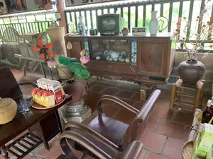 Bối cảnh là một không gian trong nhà, có thể là phòng khách hoặc khu vực trưng bày, được bài trí theo phong cách truyền thống của Việt Nam với sàn gạch đỏ. Ở giữa là một chiếc tủ chè bằng gỗ, có cửa kính, bên trong trưng bày một bộ ấm chén bằng sứ. Trên nóc tủ đặt một chiếc tivi CRT cũ hiệu 'LG', một chiếc đài radio cổ và các lọ hoa sứ.。Ở phía trước, có một bộ bàn ghế gỗ. Trên bàn có một khay tre đựng các hộp nước giải khát với chữ 'Sâm Bí Đao' và 'Trà Sâm Dứa', cùng với vài chiếc cốc thủy tinh. Bên cạnh bàn là một bình gốm lớn cắm hoa sen giả.。Không gian còn được trang trí bằng một bình gốm khác cắm cành hoa đào, gợi không khí ngày Tết. Xung quanh có các vật dụng mang đậm nét làng quê Việt Nam như một chiếc quạt điện, các loại giỏ, thúng, mủng đan bằng tre. Không có người trong ảnh, cảnh vật tĩnh lặng, mang dáng vẻ hoài cổ.。（日本語訳）。背景は、赤いタイルの床を持つ、ベトナムの伝統的なスタイルで装飾された屋内の空間で、リビングルームまたは展示エリアのようです。中央にはガラス扉の付いた木製の茶器棚があり、中には磁器の茶器一式が飾られています。棚の上には、「LG」製の古いブラウン管テレビ、アンティークのラジオ、そして磁器の花瓶が置かれています。手前には、木製のテーブルと椅子のセットがあります。テーブルの上には、「Sâm Bí Đao」と「Trà Sâm Dứa」と書かれた清涼飲料水の箱を乗せた竹製の盆と、いくつかのガラスのコップがあります。テーブルの横には、蓮の造花が生けられた大きな陶器の壺があります。空間はさらに、テト（旧正月）の雰囲気を醸し出す、桃の枝が生けられた別の陶器の壺で飾られています。周りには、扇風機、竹で編んだ様々な種類の籠やざるなど、ベトナムの田舎の趣を色濃く持つ品々があります。写真には人は写っておらず、景色は静かで、ノスタルジックな趣があります。