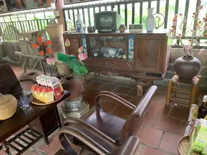 Đây là không gian bên trong một ngôi nhà hoặc khu vực nghỉ ngơi có mái che, sàn lát gạch đỏ. Không gian được bài trí theo phong cách truyền thống Việt Nam. Ở trung tâm có một chiếc tủ chè bằng gỗ, bên trong tủ kính trưng bày một bộ ấm chén trà bằng sứ. Trên nóc tủ đặt một chiếc tivi cũ hiệu LG, một chiếc đài radio cũ hiệu SONY, và hai lọ hoa sứ. Bên cạnh tủ, một bình gốm lớn đặt trên đôn đá đang cắm hoa sen. Ở góc phải, một bình gốm khác đặt trên ghế đẩu bằng tre cắm một cành hoa đào màu hồng có trang trí đèn lồng đỏ nhỏ, gợi không khí ngày Tết. Phía trước có một bộ bàn ghế gỗ. Trên bàn có một khay tre đựng các hộp nước uống có chữ "Sâm Báo", "Tâm An", vài chiếc cốc thủy tinh và một chiếc giỏ tre đan có nắp. Ở góc trái có một chiếc quạt điện màu cam hiệu AKiFan và các vật dụng làm nông bằng tre đan như nong, nia. Khung cảnh cho thấy một không gian sinh hoạt hoặc tiếp khách được trang trí công phu.。（日本語訳）。これは、家の中、あるいは屋根があり、床が赤いタイルで敷き詰められた休憩エリアの空間です。この空間はベトナムの伝統的なスタイルで装飾されています。中央には木製の茶棚があり、ガラス戸の中には磁器の茶器セットが飾られています。棚の上にはLG製の古いテレビ、SONY製の古いラジオ、そして2つの磁器の花瓶が置かれています。棚の横には、石の台座の上に置かれた大きな陶器の壺に蓮の花が生けられています。右隅には、竹製のスツールの上に置かれた別の陶器の壺に、小さな赤い提灯で飾られたピンク色の桃の花の枝が生けられており、テト（旧正月）の雰囲気を醸し出しています。手前には木製のテーブルと椅子のセットがあります。テーブルの上には、「Sâm Báo」「Tâm An」と書かれた飲み物の箱、いくつかのガラスのコップ、そして蓋付きの竹で編まれた籠が乗った竹製のお盆があります。左隅にはAKiFan製のオレンジ色の扇風機と、「ノン」や「ニア」のような竹で編まれた農具があります。この光景は、精巧に装飾された生活空間または応接空間であることを示しています。