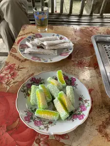 Trên một chiếc bàn có trải tấm khăn nhựa hoa văn đã cũ, có hai đĩa bánh kẹo và một cốc trà. Đĩa ở gần hơn đựng nhiều miếng bánh dẻo hai màu xanh lá cây và vàng, bên ngoài phủ vụn dừa trắng. Đĩa ở phía sau đựng các thanh kẹo được gói trong giấy bóng kính. Bên cạnh đó là một cốc thủy tinh đựng trà. Ở góc trên bên trái, có thể thấy một phần của một người đang ngồi. Cảnh tượng này mô tả một buổi thưởng thức đồ ăn nhẹ và trà, một sinh hoạt quen thuộc.。（日本語訳）。古い模様のビニール製テーブルクロスが敷かれたテーブルの上に、菓子皿が二枚と一杯のお茶がある。手前の皿には、緑色と黄色の二色の餅菓子がたくさん入っており、外側は白いココナッツフレークで覆われている。奥の皿には、セロハンで包まれた棒状のキャンディーが入っている。その隣には、お茶が入ったガラスのコップがある。この光景は、軽食とお茶を楽しむひととき、よくある日常の一コマを描写している。