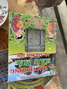 Hình ảnh chụp cận cảnh một hộp bánh đặc sản đặt trên một bề mặt trông giống như bàn gỗ. Trên vỏ hộp có các dòng chữ: 'Bánh VĂN TRƯỜNG RAU MÁ', 'Đặc Sản Sâm Sơn', và 'CƠ SỞ SẢN XUẤT BÁNH GIA TRUYỀN'. Vỏ hộp có màu xanh lá cây, được trang trí bằng hình ảnh phong cảnh núi non và hai em bé mặc trang phục truyền thống đang cầm đồng tiền vàng có chữ Hán ('吉祥如意' - Cát tường như ý). Qua một ô cửa sổ bằng nhựa trong, có thể thấy những chiếc bánh màu xanh được phủ một lớp vụn trắng, có thể là dừa. Trên hộp cũng ghi rõ thông tin sản phẩm: 'Sản phẩm: - Không sử dụng chất bảo quản. - Không màu thực phẩm. - Không đường hóa học.' và ngày sản xuất, hạn sử dụng: 'HSX: 24 6 2024', 'HSD: 30 9 202...'. Cảnh vật xung quanh bao gồm một phần của chiếc đĩa sứ có hoa văn ở bên trái.。（日本語訳）。木製のテーブルのような表面に置かれた特産品の菓子箱のクローズアップ写真。箱のパッケージには、「Bánh VĂN TRƯỜNG RAU MÁ」、「Đặc Sản Sâm Sơn」（サムソン特産）、そして「CƠ SỞ SẢN XUẤT BÁNH GIA TRUYỀN」（伝統菓子製造所）という文字が書かれています。箱は緑色で、山々の風景と、漢詩（「吉祥如意」 - 吉祥如意）が書かれた金貨を持つ伝統衣装を着た二人の子供の絵で飾られています。透明なプラスチックの窓からは、白い粉（おそらくココナッツ）がまぶされた緑色の菓子が見えます。箱には製品情報も明記されています：「製品： - 保存料不使用。- 着色料不使用。- 化学甘味料不使用。」そして製造日、賞味期限：「製造日：2024年6月24日」、「賞味期限：202...年9月30日」。周囲の背景には、左側に模様の入った磁器の皿の一部が含まれています。