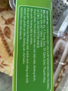 Hình ảnh chụp cận cảnh nhãn thông tin sản phẩm trên một hộp giấy màu xanh lá cây. Nội dung chi tiết trên nhãn được ghi lại như sau:。Sản xuất tại: DOANH NGHIỆP SẢN XUẤT BÁNH KẸO VĂN TRƯỜNG。ĐC: KP Bắc Kỳ, P. Trung Sơn, TP. Sầm Sơn, Thanh Hóa。ĐT: 0936 309 507 * 0963 770888。Số CBCL: 65/2017/YTTH-XNCB。Thành phần: Tinh bột củ mài, rau má, bột nếp, đường kính, đậu xanh, hương liệu tự nhiên.。Bảo quản: Nơi khô ráo thoáng mát, tránh ánh nắng mặt trời.。HDSD: Sản phẩm ăn liền, không cần chế biến.。NSX/HSD: Xem trên bao bì.。（日本語訳）。緑色の紙箱に貼られた製品情報ラベルのクローズアップ写真。ラベルに記載されている詳細な内容は以下の通りです。製造元：VAN TRUONG製菓企業。住所：タインホア省 サムソン市 チュンソン坊 バッキー街区。電話番号：0936 309 507 * 0963 770888。品質公表番号：65/2017/YTTH-XNCB。原材料：ヤマイモ澱粉、ツボクサ、もち米粉、グラニュー糖、緑豆、天然香料。保存方法：涼しく乾燥した場所に保管し、直射日光を避けてください。使用方法：調理不要、そのままお召し上がりいただけます。製造年月日/賞味期限：パッケージに記載。