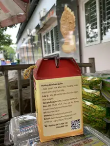 Bức ảnh chụp cận cảnh mặt sau của một hộp sản phẩm bằng bìa cứng màu đỏ và vàng, có quai xách bằng dây vàng. Hộp được đặt trên một bề mặt, có thể là một quầy hàng, ở ngoài trời. Trên nhãn của hộp có ghi chi tiết thông tin về sản phẩm và nhà sản xuất.。Nội dung trên nhãn:。- Hướng dẫn bảo quản: Bảo quản nơi khô ráo, thoáng mát, tránh ánh sáng trực tiếp. Lưu ý: Khi đã mở lọ nên cho vào ngăn mát tủ lạnh.。- Sản phẩm của: HTX Dược liệu Xứ Thanh。- Địa chỉ: Thôn Vĩnh Thọ, xã An Nông, huyện Triệu Sơn, tỉnh Thanh Hóa。- ĐT: 0947.862.678。- Website: duoclieuxuthanh.com。- Email: duoclieuxuthanh@gmail.com。- ATTP số: 162/2022/ATTP-CNĐK。- Hotline: 0878 136 136, 0966 968 736。- Có một mã QR trên nhãn.。Xung quanh là các hộp sản phẩm khác, một số có hoa văn màu xanh lá và vàng, một số được đựng trong hộp nhựa trong suốt. Phía dưới có một nhãn hiệu khác đọc được một phần là "CƠ SỞ SẢN XUẤT BÁNH KẸO GIA TRUYỀN ...ANG XUÂN ... Lộc - Thanh Hóa". Bối cảnh phía sau là một ngôi nhà có cửa sổ khung trắng và một dàn nóng điều hòa không khí, cho thấy địa điểm có thể là trước hiên nhà hoặc một cửa hàng.。（日本語訳）。赤と黄色の厚紙でできた、黄色の紐の持ち手が付いた製品箱の裏側のクローズアップ写真。箱のラベルには、製品と製造者に関する詳細情報が記載されている。ラベルの内容：。- 保管方法：乾燥した涼しい場所に保管し、直射日光を避けてください。注意：瓶を開封した後は、冷蔵庫の冷蔵室に入れてください。- 製品提供元：HTX Dược liệu Xứ Thanh（スータン薬草協同組合）。- 住所：タインホア省、トリウソン県、アンノン社、ヴィントー村。- 電話番号：0947.862.678。- ウェブサイト：duoclieuxuthanh.com。- Eメール：duoclieuxuthanh@gmail.com。- 食品安全証明書番号：162/2022/ATTP-CNĐK。- ホットライン：0878 136 136, 0966 968 736。- ラベルにはQRコードがあります。周りには他の製品箱があり、緑と黄色の模様が入ったものや、透明なプラスチックの箱に入ったものもある。下の方には、部分的に「CƠ SỞ SẢN XUẤT BÁNH KẸO GIA TRUYỀN ...ANG XUÂN ... Lộc - Thanh Hóa」（家伝菓子製造所...アン・スアン...ロック - タインホア）と読める別のブランドラベルがある。背景には白い窓枠の家とエアコンの室外機があり、場所は家の軒先か店舗の前である可能性を示している。