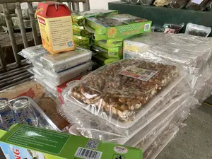 Bối cảnh là một quầy hàng trưng bày các sản phẩm đặc sản địa phương để bán, có thể là một cửa hàng hoặc gian hàng ở chợ. Các sản phẩm được xếp chồng lên nhau. Ở phía trước là các gói kẹo lạc được đóng gói trong khay xốp và bọc nilon. Nhãn trên sản phẩm ghi "ĐẶC SẢN SỨ THANH KẸO CU ĐƠ SẦM SƠN" và có logo OCOP. Phía sau là các hộp sản phẩm khác. Một hộp màu đỏ và vàng có ghi: "SÂM BẢO Mật Ong". Thông tin chi tiết trên hộp: "Hướng dẫn bảo quản: - Bảo quản nơi khô ráo, thoáng mát, tránh ánh sáng trực tiếp. Lưu ý: Khi đã mở lọ nên cho vào ngăn mát tủ lạnh. Sản phẩm của: HTX Dược liệu Xứ Thanh. Địa chỉ: Thôn Vĩnh Thọ, xã An Nông, huyện Triệu Sơn, tỉnh Thanh Hóa. ĐT: 0947.862.678. Website: duoclieuxuthanh.com. Email: duoclieuxuthanh@gmail.com. ATTP số: 162/2022/ATTP-CNĐK. Hotline: 0878 136 136, 0966 968 736". Bên cạnh đó là các hộp màu xanh lá cây có chữ "Bánh Đặc Sản Sầm Sơn RAU MÁ". Thông tin trên hộp: "Sản xuất tại: DOANH NGHIỆP SẢN XUẤT BÁNH KẸO VĂN TRƯỜNG. ĐC: KP Bắc Kỳ, P. Trung Sơn, TP. Sầm Sơn, Thanh Hóa. Thành phần: Tinh bột củ mài, rau má, bột nếp, đường kính, đậu xanh, hương liệu tự nhiên. Bảo quản: Nơi khô ráo thoáng mát, tránh ánh nắng mặt trời. HDSD: Sản phẩm ăn liền, không cần chế biến. NSX/HSD: Xem trên bao bì". Hộp này cũng có logo "OCOP THANH HÓA". Ngoài ra, ở bên trái còn có một vài chiếc cốc thủy tinh đặt trên khay kim loại. Hoạt động chính là trưng bày và bán các mặt hàng đặc sản này.。（日本語訳）。背景は、地元の特産品を販売するために陳列している売店で、店舗か市場のブースのようです。商品は積み重ねられています。手前には、発泡スチロールのトレイに詰められ、ナイロンで包装されたピーナッツ菓子があります。商品のラベルには「タイン省特産 クドー菓子 サムソン」と書かれており、OCOPのロゴがあります。後ろには他の商品の箱があります。赤と黄色の箱には「サムバオ 蜂蜜」と書かれています。箱の詳細情報：「保存方法：- 乾燥した涼しい場所に保管し、直射日光を避けてください。注意：瓶を開封した後は、冷蔵庫の冷暗所に入れてください。製造元：タイン省薬草協同組合。住所：タインホア省、トリウソン県、アンノン社、ヴィントー村。電話：0947.862.678。ウェブサイト：duoclieuxuthanh.com。メール：duoclieuxuthanh@gmail.com。食品安全衛生番号：162/2022/ATTP-CNĐK。ホットライン：0878 136 136、0966 968 736」。その隣には、「サムソン特産 ツボクサケーキ」と書かれた緑色の箱があります。箱の情報：「製造元：ヴァンチュオン製菓企業。住所：タインホア省、サムソン市、チュンソン区、バックキー街区。原材料：ヤマイモ澱粉、ツボクサ、もち米粉、グラニュー糖、緑豆、天然香料。保存方法：乾燥した涼しい場所に保管し、直射日光を避けてください。使用方法：調理不要のインスタント製品です。製造年月日/賞味期限：パッケージをご覧ください」。この箱にも「OCOP THANH HÓA」のロゴがあります。また、左側には金属製のトレイの上にいくつかのガラスのコップが置かれています。主な活動は、これらの特産品の陳列と販売です。