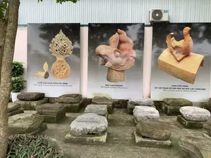 Đây là một khu trưng bày hiện vật ngoài trời, có thể là một phần của di tích lịch sử hoặc bảo tàng. Ở tiền cảnh, nhiều khối đá lớn, có thể là chân cột đá của các công trình kiến trúc cổ, được đặt trên các bệ bê tông. Phía sau là một bức tường có gắn ba tấm pano lớn in hình các hiện vật khảo cổ bằng đất nung. Trên tường, phía trên bên phải, có một cục nóng điều hòa không khí hiệu Casper. Bên trái có một thân cây và một vài cây cảnh nhỏ.。Nội dung chữ viết trên các tấm pano được ghi lại như sau:。- Tấm pano bên trái: "LÁ ĐỀ CÂN CHẠM RỒNG ĐẤT NUNG (khai quật ở chính điện Kinh thành Tây Đô)". Pano này trưng bày hình ảnh một vật trang trí kiến trúc hình lá đề bằng đất nung.。- Tấm pano ở giữa: "ĐẦU CHIM PHƯỢNG (Khai quật tại Đàn tế Nam Giao)". Pano này trưng bày hình ảnh một đầu tượng chim phượng bằng đất nung.。- Tấm pano bên phải: "CHIM UYÊN ƯƠNG VẬT LIỆU TRANG TRÍ KIẾN TRÚC BỜ NÓC CÁC CUNG ĐIỆN (Hiện vật khai quật tại Thành Nhà Hồ)". Pano này trưng bày hình ảnh một vật liệu trang trí mái nhà hình chim uyên ương.。（日本語訳）。これは屋外の遺物展示エリアで、歴史遺跡または博物館の一部である可能性があります。前景には、古代建築物の石柱の礎石と思われる多くの大きな石塊が、コンクリートの台座の上に置かれています。後ろには、テラコッタ製の考古学的遺物の写真が印刷された3枚の大きなパネルが取り付けられた壁があります。壁の右上部には、Casper（キャスパー）ブランドのエアコンの室外機が1台あります。左側には木の幹と、いくつかの小さな観葉植物があります。各パネルに書かれている内容は以下の通りです。- 左側のパネル：「テラコッタ製 龍彫刻の菩提樹葉形装飾（西都城の正殿で発掘）」。このパネルは、テラコッタ製の菩提樹の葉の形をした建築装飾物の画像を展示しています。- 中央のパネル：「鳳凰の頭部（南郊壇にて発掘）」。このパネルは、テラコッタ製の鳳凰の像の頭部の画像を展示しています。- 右側のパネル：「鴛鴦 宮殿の屋根の棟の建築装飾材（胡朝の城にて発掘された遺物）」。このパネルは、鴛鴦の形をした屋根装飾材の画像を展示しています。