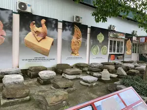 Bức ảnh chụp một khu trưng bày hiện vật khảo cổ ngoài trời, có khả năng là tại khu di tích Thành Nhà Hồ. Dưới mặt đất, có rất nhiều khối đá cổ, có vẻ là các chân tảng cột, được đặt trên các bệ bê tông. Phía sau là bức tường của một tòa nhà, có treo nhiều tấm pano lớn in hình các hiện vật khảo cổ đã được khai quật.。Nội dung chữ viết trên các tấm pano bao gồm:。- Tấm pano bên trái có hình một con chim bằng đất nung và ghi: "CHIM UYÊN ƯƠNG, VẬT LIỆU TRANG TRÍ KIẾN TRÚC BỜ NÓC CÁC CUNG ĐIỆN (Hiện vật khai quật tại Thành Nhà Hồ)".。- Tấm pano ở giữa có hình một lá đề bằng đất nung chạm khắc rồng và ghi: "LÁ ĐỀ LỆCH CHẠM RỒNG ĐẤT NUNG (Khai quật ở chính diện Kinh thành Tây Đô)".。- Tấm pano tiếp theo có hình các lá đề men vàng và ghi: "LÁ ĐỀ CÂN MEN VÀNG (Khai quật ở chính điện Kinh thành Tây Đô)".。- Các hình ảnh khác trên tường còn có chú thích "NGÓI ÂM DƯƠNG", "GẠCH LÁT NỀN TRANG TRÍ RỒNG", và "LÁ ĐỀ CÂN CHẠM RỒNG ĐẤT NUNG".。Trên tường tòa nhà có gắn nhiều cục nóng điều hòa và một biển hiệu UNESCO. Cảnh vật có cây xanh.。（日本語訳）。写真は、屋外の考古学遺物展示エリアを写したもので、おそらく胡朝の城塞遺跡にあるものだと思われます。地面には、コンクリートの台座の上に置かれた、柱の礎石と思われる古代の石の塊が多数あります。後方には建物の壁があり、そこには発掘された考古学遺物の写真が印刷された大きなパネルが多数掛けられています。パネルに書かれている文字の内容は以下の通りです。- 左側のパネルには、テラコッタの鳥の絵があり、「オシドリ、宮殿の屋根の棟の建築装飾材（胡朝の城塞で出土した遺物）」と書かれています。- 中央のパネルには、龍が彫られたテラコッタの菩提樹の葉の絵があり、「龍が彫られたテラコッタの非対称な菩提樹の葉（西都城の正面で発掘）」と書かれています。- 次のパネルには、黄釉の菩提樹の葉の絵があり、「黄釉の対称な菩提樹の葉（西都城の正殿で発掘）」と書かれています。- 壁にある他の写真には、「陰陽瓦」、「龍の装飾が施された床タイル」、および「龍が彫られたテラコッタの対称な菩提樹の葉」というキャプションも付いています。建物の壁には、多くのエアコンの室外機とユネスコの看板が取り付けられています。風景には緑の木々があります。