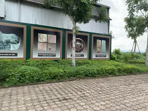 Bối cảnh là một khu vực trưng bày ngoài trời, có thể là tại một khu di tích lịch sử. Dọc theo một lối đi lát gạch hình bát giác là một dãy các tấm biển thông tin lớn màu xanh lá cây. Trên các tấm biển này có trưng bày hình ảnh các hiện vật khảo cổ. Phía sau các tấm biển là một tòa nhà có tường bằng tôn và cây cối.。Nội dung chữ viết trên các tấm biển, từ trái sang phải:。1. Biển thứ nhất (bị che một phần): Hình một pho tượng đá với dòng chữ "...Ử ĐÁ CHÙA DU ANH".。2. Biển thứ hai: Hình bốn viên gạch trang trí với dòng chữ "GẠCH TRANG TRÍ HOA CHANH" và bên dưới là "KHAI QUẬT TẠI NỘI THÀNH".。3. Biển thứ ba (bị che một phần bởi thân cây): Hình một chiếc bát gốm với dòng chữ "...ÁT MEN NÂU" và bên dưới là "TẠI ĐÀN TẾ NAM GIAO - TÂY ĐÔ".。4. Biển thứ tư: Hình hai viên gạch có khắc chữ với dòng chữ "GẠCH BÌA CÓ MINH VĂN" và bên dưới là "KHAI QUẬT TẠI THÀNH NHÀ HỒ".。（日本語訳）。背景は屋外の展示エリアで、歴史遺跡の敷地内である可能性があります。八角形のタイルが敷かれた通路に沿って、緑色の大きな案内板の列があります。これらの案内板には、考古学的な遺物の写真が展示されています。案内板の後ろには、トタンの壁の建物と木々があります。案内板に書かれた文字の内容、左から右へ：。1. 1枚目の案内板（一部が隠れている）：石像の写真と「...Ử ĐÁ CHÙA DU ANH」という文字。2. 2枚目の案内板：4つの装飾レンガの写真と「GẠCH TRANG TRÍ HOA CHANH」という文字、そしてその下には「KHAI QUẬT TẠI NỘI THÀNH」。3. 3枚目の案内板（木の幹で一部が隠れている）：陶器の碗の写真と「...ÁT MEN NÂU」という文字、その下には「TẠI ĐÀN TẾ NAM GIAO - TÂY ĐÔ」。4. 4枚目の案内板：文字が刻まれた2つのレンガの写真と「GẠCH BÌA CÓ MINH VĂN」という文字、その下には「KHAI QUẬT TẠI THÀNH NHÀ HỒ」。