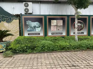 Bối cảnh là một không gian ngoài trời, có thể là khu vực trưng bày của một di tích lịch sử. Trên một bức tường tôn, có một dãy các bảng thông tin lớn màu xanh lá cây trưng bày hình ảnh các hiện vật. Phía trước là một hàng cây bụi và vỉa hè lát gạch block hình lục giác.。Nội dung chữ viết trên các bảng thông tin:。1. Bảng bên trái có hình một con sư tử đá với chú thích: 'SƯ TỬ ĐÁ CHÙA DU ANH'.。2. Bảng ở giữa có hình bốn viên gạch trang trí với chú thích: 'GẠCH TRANG TRÍ HOA CHANH' và 'KHAI QUẬT TẠI NỘI THÀNH'.。3. Bảng bên phải, bị một thân cây che khuất một phần, có hình một chiếc bát hoặc đĩa gốm và chú thích có thể đọc được là '...T MEN NÂU' và 'ĐÀN TẾ NAM GIAO - TÂY ĐÔ'.。Các vật thể khác trong ảnh bao gồm một cục nóng điều hòa không khí hiệu LG gắn trên tường, một tấm trang trí lớn bên trái có họa tiết rồng màu trắng trên nền màu be, và một chậu cây cảnh lớn màu xanh ngọc.。（日本語訳）。背景は屋外の空間で、史跡の展示エリアである可能性があります。トタンの壁には、遺物の写真を展示している緑色の大きな案内板が並んでいます。手前には一列の低木と、六角形のブロックタイルで舗装された歩道があります。案内板に書かれている文字の内容：。1. 左側のパネルには石獅子の写真があり、「SƯ TỬ ĐÁ CHÙA DU ANH」というキャプションが付いています。2. 中央のパネルには4枚の装飾タイルの写真があり、「GẠCH TRANG TRÍ HOA CHANH」と「KHAI QUẬT TẠI NỘI THÀNH」というキャプションが付いています。3. 右側のパネルは、木の幹で一部が隠れており、陶器の碗または皿の写真と、読み取れるキャプションとして「...T MEN NÂU」および「ĐÀN TẾ NAM GIAO - TÂY ĐÔ」があります。写真にあるその他の物体には、壁に取り付けられたLG製のエアコン室外機、ベージュの背景に白い龍の模様がある左側の大きな装飾パネル、そして青緑色の大きな観葉植物の鉢が含まれます。