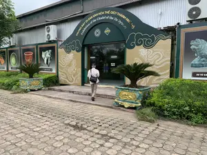 Bối cảnh là lối vào một tòa nhà, có thể là một khu trưng bày hoặc bảo tàng. Phía trên lối vào hình vòm có dòng chữ lớn màu vàng: "TRƯNG BÀY DI SẢN VĂN HÓA THẾ GIỚI THÀNH NHÀ HỒ" và bên dưới là dòng chữ tiếng Anh "Exhibition of the Citadel of the Ho Dynasty". Trên cổng còn có logo Di sản Thế giới của UNESCO và logo Thành nhà Hồ. Một người đàn ông lớn tuổi mặc áo sơ mi, đội mũ và đeo túi chéo đang đi lên bậc thang để vào bên trong qua cửa kính. Trên cửa kính có ghi "ĐẨY VÀO PUSH IN". Hai bên lối vào có hai chậu cây cảnh lớn, trang trí hoa văn màu vàng, trồng cây vạn tuế. Dọc theo các bức tường bên ngoài là các tấm pano lớn trưng bày hình ảnh các hiện vật khảo cổ, với các chú thích như "BÁT MEN XANH", "ĐẦU RỒNG ĐÁ THỜI TRẦN - HỒ", và "SƯ TỬ ĐÁ". Sân trước được lát bằng gạch block hình lục giác. Tòa nhà có tường và mái bằng tôn, bên trên có lắp đặt các dàn nóng của máy điều hòa không khí.。（日本語訳）。背景は、ある建物の入り口で、展示エリアか博物館のようです。アーチ型の入り口の上には、大きな黄色い文字で「TRƯNG BÀY DI SẢN VĂN HÓA THẾ GIỚI THÀNH NHÀ HỒ」と書かれており、その下には英語で「Exhibition of the Citadel of the Ho Dynasty」と書かれています。門の上には、ユネスコ世界遺産のロゴと胡朝の城塞のロゴもあります。シャツを着て、帽子をかぶり、ショルダーバッグを斜め掛けした年配の男性が、ガラスのドアから中に入るために階段を上っています。ガラスのドアには「ĐẨY VÀO PUSH IN」と書かれています。入り口の両側には、黄色い模様で装飾された大きな観葉植物の鉢が2つあり、ソテツが植えられています。外壁に沿って、考古学的な遺物の写真が展示された大きなパネルがあり、「BÁT MEN XANH」、「ĐẦU RỒNG ĐÁ THỜI TRẦN - HỒ」、「SƯ TỬ ĐÁ」などのキャプションが付いています。前庭は六角形のブロックタイルで舗装されています。建物はトタンの壁と屋根で、その上にはエアコンの室外機が設置されています。