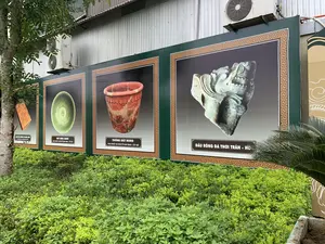 Bối cảnh là một khu vực trưng bày ngoài trời, có các tấm pano thông tin lớn màu xanh lá cây được dựng lên trước một tòa nhà có tường bằng tôn. Phía trước các tấm pano là một luống cây bụi xanh tốt. Các tấm pano này trưng bày hình ảnh và thông tin về các cổ vật lịch sử được khai quật tại khu vực Thành nhà Hồ. Nội dung chữ viết trên các tấm pano từ trái sang phải như sau:。1. Tấm pano thứ nhất: "GẠCH BÌA KHÔNG CÓ MINH VĂN", "KHAI QUẬT TẠI THÀNH NHÀ HỒ". Hình ảnh là một viên gạch.。2. Tấm pano thứ hai: "BÁT MEN XANH", "KHAI QUẬT TẠI ĐÀN TẾ NAM GIAO - TÂY ĐÔ". Hình ảnh là một chiếc bát tráng men màu xanh.。3. Tấm pano thứ ba: "THỐNG ĐẤT NUNG", "KHAI QUẬT TẠI ĐÀN TẾ NAM GIAO - TÂY ĐÔ". Hình ảnh là một chiếc vại bằng đất nung.。4. Tấm pano thứ tư: "ĐẦU RỒNG ĐÁ THỜI TRẦN - HỒ". Hình ảnh là một đầu rồng bằng đá. Phía sau các tấm pano có thể thấy một phần của tòa nhà và các cục nóng điều hòa không khí.。（日本語訳）。背景は屋外の展示エリアで、トタン壁の建物の前に大きな緑色の情報パネルが設置されています。パネルの前には、青々とした低木の植え込みがあります。これらのパネルは、胡朝の城の地域で発掘された歴史的な遺物に関する写真と情報を展示しています。パネルに書かれている内容は、左から右へ以下の通りです。1. 1枚目のパネル：「銘文のない化粧煉瓦」、「胡朝の城にて発掘」。写真は煉瓦です。2. 2枚目のパネル：「青磁の碗」、「南郊壇 - 西都にて発掘」。写真は青い釉薬のかかった碗です。3. 3枚目のパネル：「土製の壺」、「南郊壇 - 西都にて発掘」。写真は土製の甕です。4. 4枚目のパネル：「陳-胡朝時代の石の龍の頭」。写真は石でできた龍の頭です。パネルの後ろには、建物の一部とエアコンの室外機が見えます。