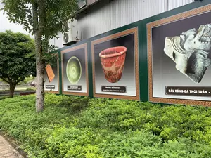 Bức ảnh chụp một không gian trưng bày ngoài trời, có thể là tại một khu di tích lịch sử. Dọc theo một bức tường tôn của một tòa nhà là một dãy các tấm pano lớn màu xanh lá cây, trên đó có in hình ảnh các hiện vật khảo cổ cùng với chú thích. Phía trước các tấm pano là một thảm cây bụi xanh tươi và một cái cây. Gắn trên tường tòa nhà có một cục nóng điều hòa. Ở phía xa, có thể thấy một người đang đi xe máy.。Nội dung chữ viết trên các tấm pano bao gồm:。- Tấm pano 1: "GẠCH BIA KHÔNG CÓ MINH VĂN", "KHAI QUẬT TẠI THÀNH NHÀ HỒ".。- Tấm pano 2: "BÁT MEN XANH", "KHAI QUẬT TẠI ĐÀN TẾ NAM GIAO - TÂY ĐÔ".。- Tấm pano 3: "THỐNG ĐẤT NUNG", "KHAI QUẬT TẠI ĐÀN TẾ NAM GIAO - TÂY ĐÔ".。- Tấm pano 4: "ĐẦU RỒNG ĐÁ THỜI TRẦN -".。Các hiện vật được trưng bày qua hình ảnh là gạch bia, bát men xanh, thống đất nung và đầu rồng bằng đá.。（日本語訳）。写真は、おそらく史跡にある屋外展示スペースを写しています。建物のトタンの壁に沿って、大きな緑色のパネルが並んでおり、そこには考古学的な遺物の写真と説明文が印刷されています。パネルの前には、青々とした低木の茂みと一本の木があります。建物の壁にはエアコンの室外機が取り付けられています。遠くには、バイクに乗っている人が見えます。パネルに書かれている文字の内容は以下の通りです。- パネル1：「銘文のないレンガ碑」、「胡朝の城跡にて発掘」。- パネル2：「青磁碗」、「南郊壇 - 西都にて発掘」。- パネル3：「土器の壺」、「南郊壇 - 西都にて発掘」。- パネル4：「陳朝時代の石の龍の頭 -」。写真で展示されている遺物は、レンガ碑、青磁碗、土器の壺、そして石の龍の頭です。