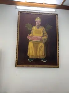 Bức ảnh chụp một bức tranh sơn dầu được treo trên tường trắng trong một căn phòng. Bức tranh được lồng trong một khung gỗ mạ vàng có hoa văn trang trí. Phía trên bức tranh có một chiếc đèn tuýp huỳnh quang đang phát sáng.。Nội dung của bức tranh là chân dung của một nhân vật lịch sử, có thể là một vị vua hoặc một vị quan lớn của triều đại phong kiến Việt Nam. Người đàn ông trong tranh có râu dài, đội mũ miện và mặc một chiếc áo bào dài màu vàng. Ông đang ngồi trên một chiếc ghế trông giống như ngai vàng, với phần tựa lưng có chạm khắc hình đầu rồng màu xanh lục. Hai tay ông đang cầm một cuộn giấy màu hồng, có vẻ là một chiếu chỉ hoặc văn thư, trên đó có các ký tự nhưng không thể đọc rõ. Ông đi một đôi hài màu sẫm.。（日本語訳）。写真は、部屋の白い壁に掛けられた一枚の油絵を写しています。その絵画は、装飾的な模様が施された金メッキの木製フレームに収められています。絵画の上には、光を放っている蛍光灯があります。絵画の内容は、歴史上の人物の肖像画で、おそらくベトナムの封建王朝の王か高官でしょう。絵の中の男性は長い髭を生やし、冠をかぶり、黄色の長い袍（ほう）を着ています。彼の両手はピンク色の巻物を握っており、それは詔勅か公文書のようですが、そこに書かれている文字ははっきりと読み取れません。彼は暗い色の履物を履いています。