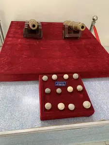 Bên trong một không gian trưng bày, có thể là bảo tàng, có các hiện vật lịch sử. Trên một bục được phủ vải nhung đỏ là hai mô hình súng thần công cỡ nhỏ bằng đồng, đặt trên giá đỡ bằng gỗ. Phía trước, trên một bục thấp hơn cũng phủ nhung đỏ, là một bộ sưu tập các viên đạn súng thần công bằng đá. Ở giữa các viên đạn có một tấm biển nhỏ màu xanh dương với dòng chữ trắng: "KHÔNG SỜ HIỆN VẬT".。（日本語訳）。展示空間、おそらくは博物館の内部には、歴史的な遺物があります。赤いビロードの布で覆われた台座の上には、木製の架台に置かれた、銅製の小さな大砲の模型が2つあります。手前には、同じく赤いビロードで覆われた低い台座の上に、石製の大砲の弾のコレクションがあります。弾の中央には、「展示物に触れないでください」と白い文字で書かれた小さな青い札があります。