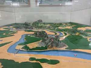 Đây là hình ảnh một sa bàn địa hình được đặt trong một tủ kính, có khả năng được trưng bày tại một bảo tàng hoặc khu di tích. Sa bàn mô phỏng một cách chi tiết cảnh quan địa lý bao gồm các con sông được sơn màu xanh lam, các vùng đồng bằng hoặc ruộng lúa màu xanh lá, và các vùng đất màu nâu vàng. Nổi bật trên sa bàn là các mô hình núi đá có cây cối, một công trình kiến trúc lớn trông giống như một thành cổ hoặc pháo đài, và một cây cầu nhỏ bắc qua sông. Rải rác trên sa bàn là các biển báo nhỏ màu đỏ có ghi số màu trắng, dùng để đánh dấu các vị trí cụ thể. Các số có thể đọc được bao gồm: 26, 27, 21, 15, 12, 10, 08, 03, 16. Không có hoạt động của con người, đây là một vật trưng bày tĩnh. Phản chiếu trên mặt kính là ánh đèn và một phần của căn phòng trưng bày.。（日本語訳）。これは、ガラスケースの中に置かれた地形ジオラマの画像で、博物館または史跡で展示されている可能性があります。ジオラマは、青色に塗られた川、緑色の平野または水田、そして黄褐色の土地を含む地理的景観を詳細に模倣しています。ジオラマには、特定の場所を印すために使われる、白い数字が書かれた小さな赤い標識が点在しています。読み取れる数字には、26、27、21、15、12、10、08、03、16が含まれます。人間の活動はなく、これは静的な展示物です。ガラス面には照明の光と展示室の一部が反射しています。