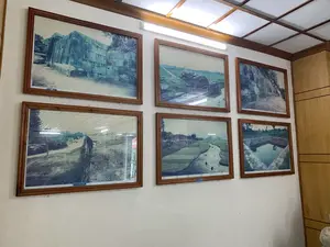 Bức ảnh chụp một bức tường bên trong một căn phòng, có khả năng là phòng trưng bày của một bảo tàng hoặc khu di tích. Trên bức tường màu trắng, có sáu bức ảnh tư liệu được đóng khung gỗ màu nâu, sắp xếp thành hai hàng. Các bức ảnh này ghi lại hình ảnh của một công trình thành cổ bằng đá, có thể là Thành nhà Hồ.。Nội dung các bức ảnh bao gồm:。- Các bức tường thành và cổng thành được xây dựng từ những khối đá lớn.。- Một cấu trúc vòm đá bị hư hại nằm giữa cánh đồng.。- Một con sông hoặc hào nước uốn lượn quanh các thửa ruộng lúa, có người đang tắm và đàn trâu bò đang gặm cỏ gần tường thành.。- Một cái giếng cổ hình vuông được kè đá cẩn thận.。- Con đường đất chạy dọc theo chân tường thành.。Dưới một vài bức ảnh có các tấm biển chú thích nhỏ màu xanh. Một trong số đó có thể đọc được dòng chữ: "Kết cấu tường phía Đông Nam" và "The southeast Wall's structure". Các chú thích khác không thể đọc rõ do chất lượng ảnh.。（日本語訳）。写真は部屋の中の壁を写したもので、博物館または史跡の展示室である可能性があります。白い壁には、茶色の木製フレームに入れられた6枚の資料写真が2列に並べられています。これらの写真は、石造りの古い城塞、おそらく胡朝の城塞の画像を記録したものです。写真の内容は以下の通りです。- 大きな石のブロックで建てられた城壁と城門。- 野原の真ん中にある損傷した石造りのアーチ構造物。- 水田の周りを蛇行する川または堀、水浴びをしている人、城壁の近くで草を食む水牛の群れ。- 丁寧に石で組まれた四角い古井戸。- 城壁の麓に沿って走る土の道。いくつかの写真の下には、小さな青いキャプションプレートがあります。そのうちの一つには、「南東壁の構造」と「The southeast Wall's structure」という文字が読み取れます。他のキャプションは写真の画質のため、はっきりと読み取れません。