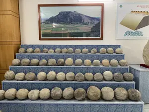 Bức ảnh chụp một không gian trưng bày trong nhà, có thể là một bảo tàng hoặc khu di tích. Các hiện vật chính là hàng chục viên đạn đá hình cầu, được sắp xếp ngay ngắn thành nhiều hàng trên các kệ bậc thang. Các kệ này được trang trí bằng hoa văn màu xanh và trắng. Phía sau khu trưng bày, trên tường có treo một bức tranh lớn trong khung gỗ, chụp phong cảnh một ngọn núi đá và những cánh đồng bên dưới. Bên phải bức tranh là một bảng thông tin có tiêu đề "MÔ HÌNH GIÁ ĐỊNH" và dòng chữ tiếng Anh "MODEL OF ST...". Bảng này có một sơ đồ minh họa một bức tường thành và các máy bắn đá, cho thấy các viên đá này là đạn dùng cho vũ khí thời xưa. Phía bên trái của căn phòng là một chiếc tủ gỗ.。（日本語訳）。写真は屋内の展示スペースを写しており、博物館または史跡の可能性があります。主な展示物は数十個の球形の石弾で、階段状の棚の上に数列にわたって整然と並べられています。これらの棚は青と白の模様で装飾されています。展示エリアの後ろの壁には、木製の額縁に入った大きな絵画が掛けられており、岩山とその下の田畑の風景が写されています。絵画の右側には、「MÔ HÌNH GIÁ ĐỊNH」というタイトルと「MODEL OF ST...」という英語の文字が書かれた案内板があります。この案内板には城壁と投石機を描いた図があり、これらの石が昔の兵器に使われた弾薬であることを示しています。部屋の左側には木製の戸棚があります。